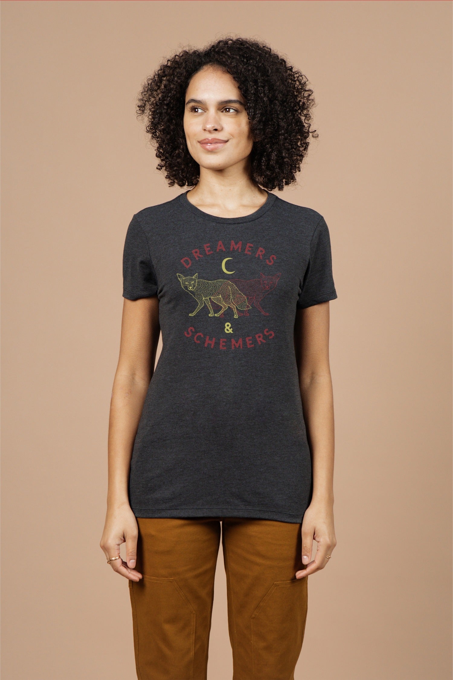 A woman with curly hair wears the Bridge & Burn Women's Dreamers & Schemers Tee in Black, featuring a fox, crescent moon, and red-yellow "DREAMERS & SCHEMERS" graphic. She stands against a beige background, looking slightly to the side.