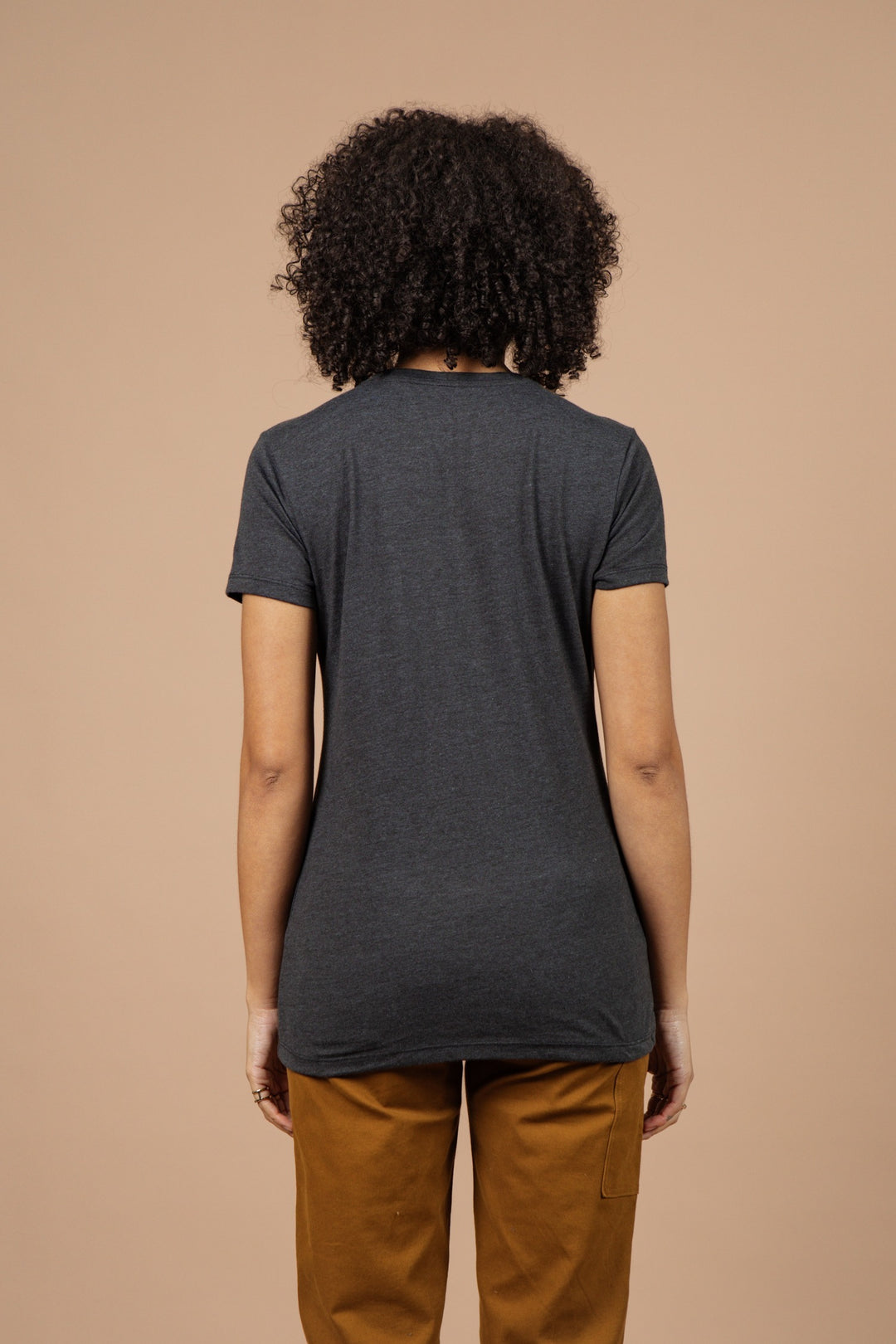 Person with curly hair wearing the Bridge & Burn Women's Dreamers & Schemers Tee in black and brown pants, standing with back to the camera against a plain beige background.