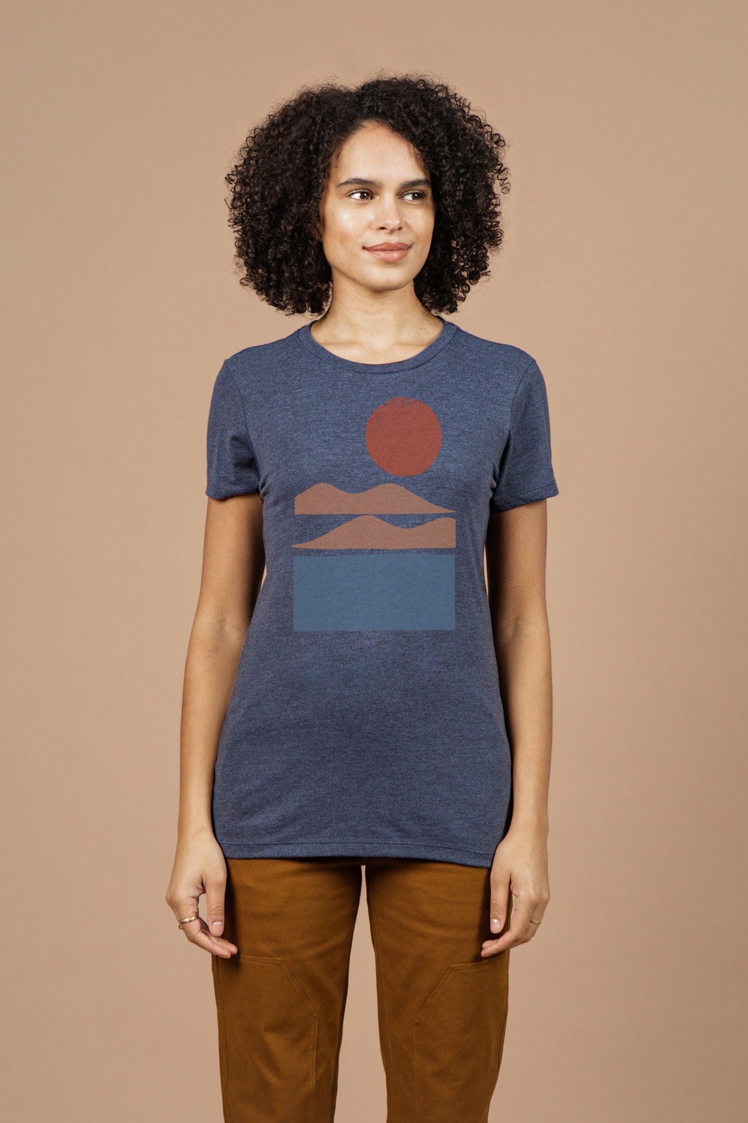A person with curly hair wears the Bridge & Burn Women's Alpenglow Tee in navy, featuring a geometric landscape design. They stand against a beige background, smiling gently to the side.