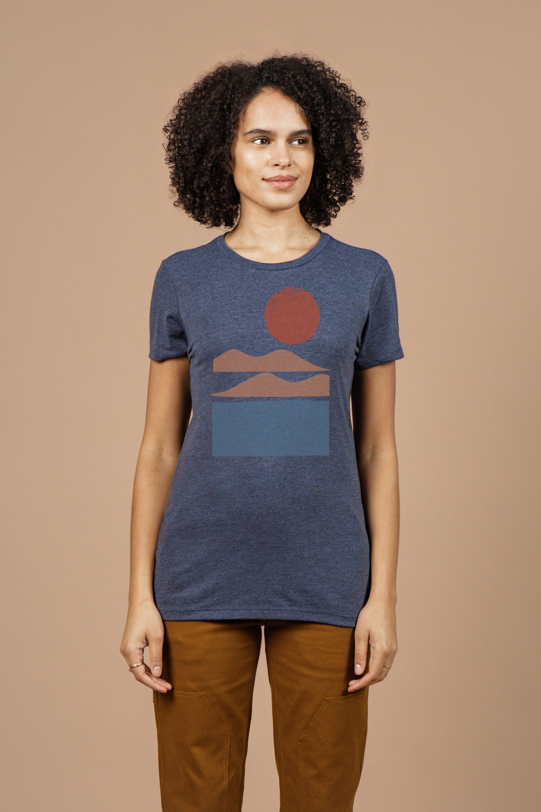 A person with curly hair wears the Bridge & Burn Women's Alpenglow Tee in navy, featuring a geometric landscape design. They stand against a beige background, smiling gently to the side.