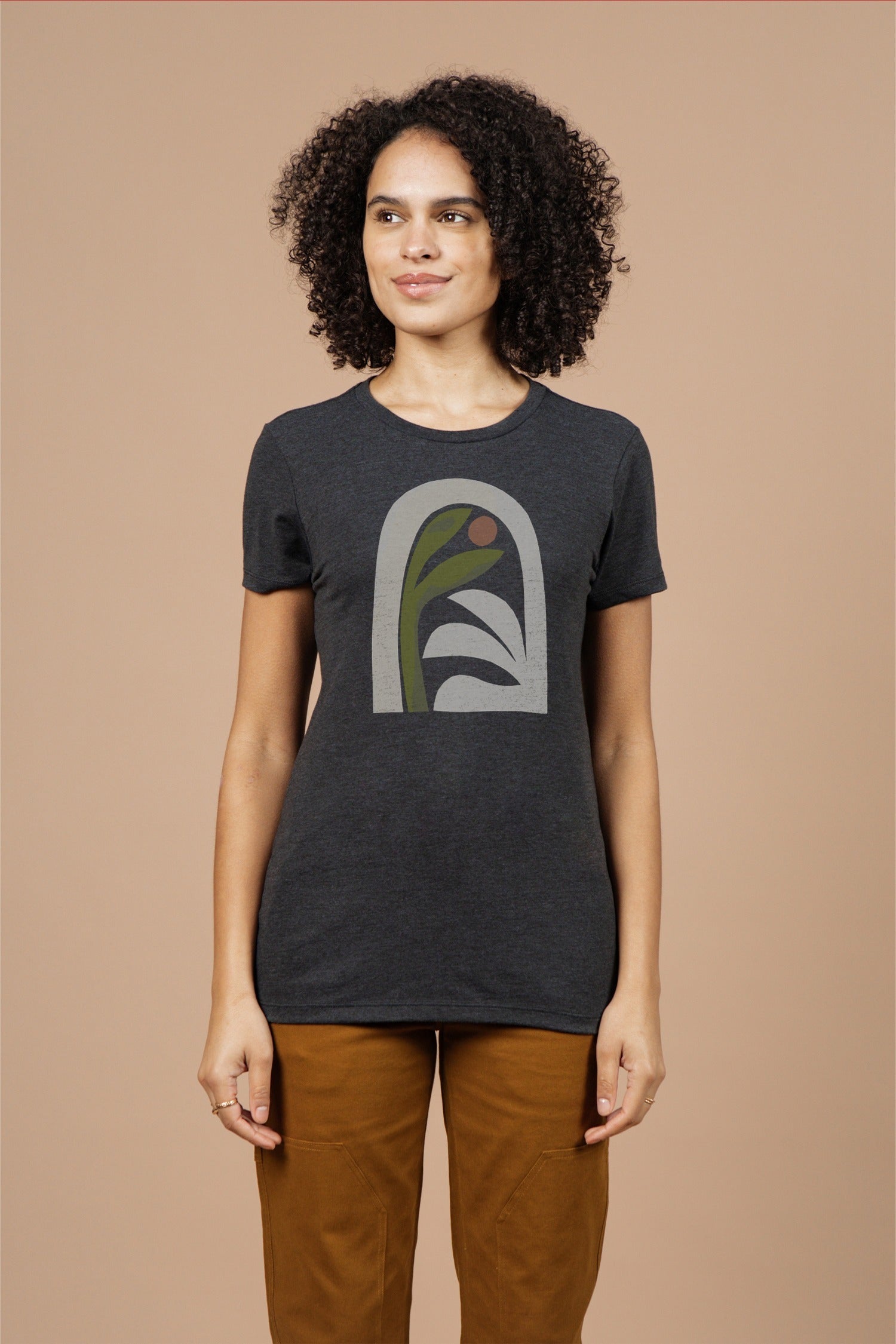 A woman with curly hair wears the Bridge & Burn Women's Arch Tee in Charcoal and brown pants, standing against a plain beige background.