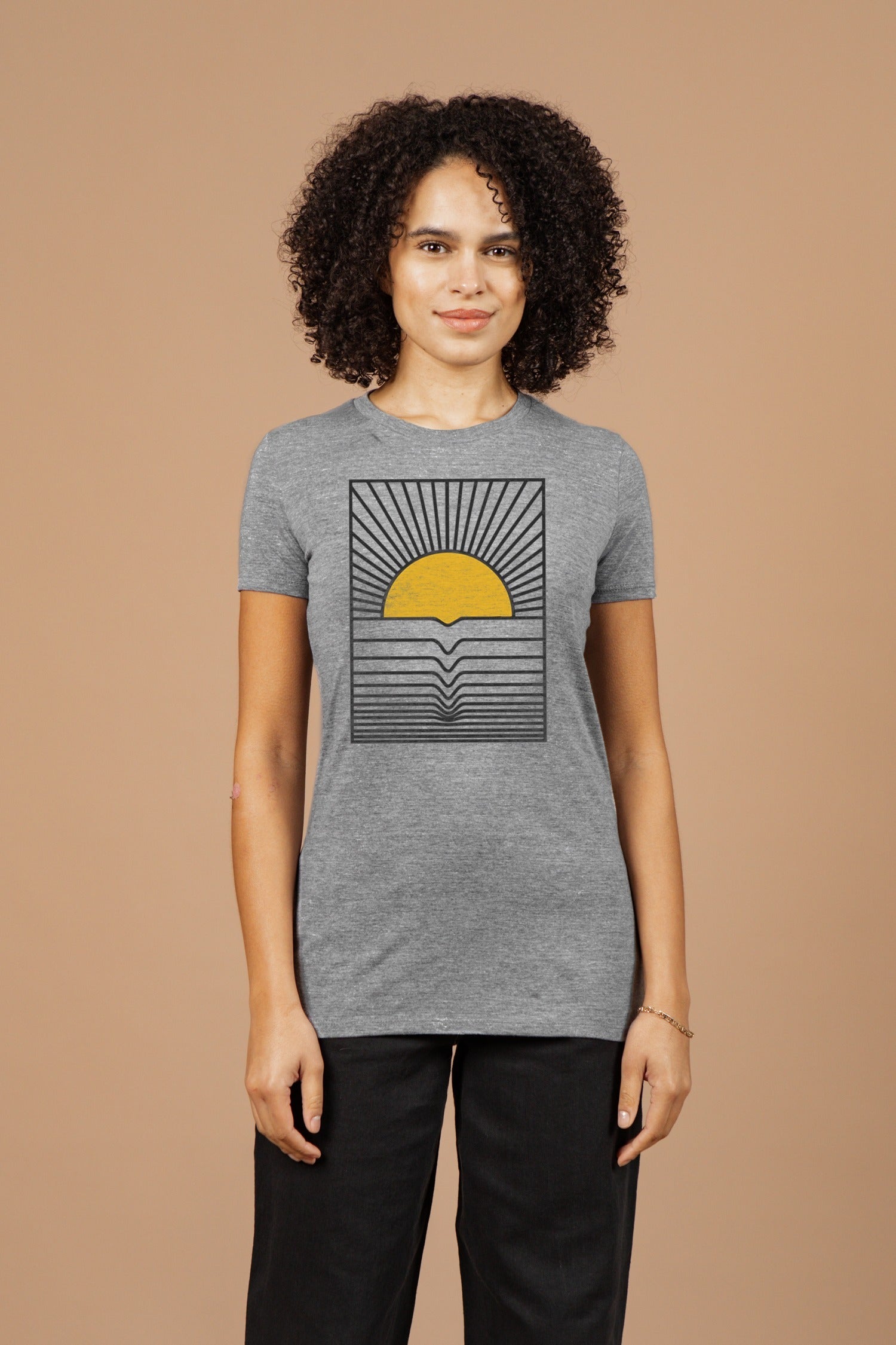 A person with curly hair wears the Bridge & Burn Women's Cresting Wave Tee in grey, featuring a yellow sun setting over stylized waves with radiating black lines, against a plain beige background.