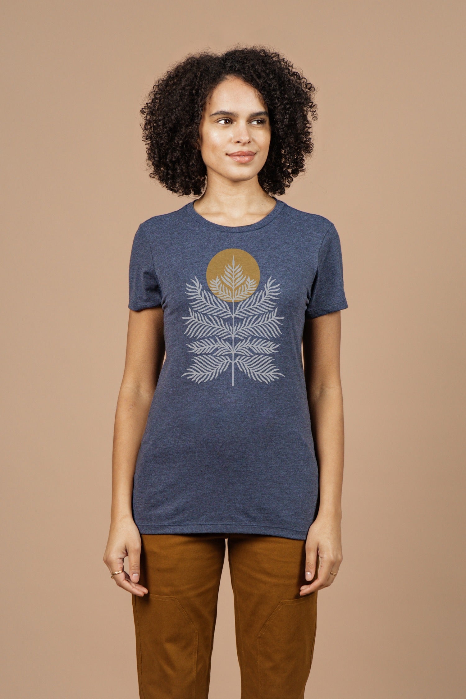 A woman with curly hair wears the Bridge & Burn Women's Peace In Plants Tee in navy, featuring a white leaf and gold circle design, paired with brown pants, standing against a plain beige background.