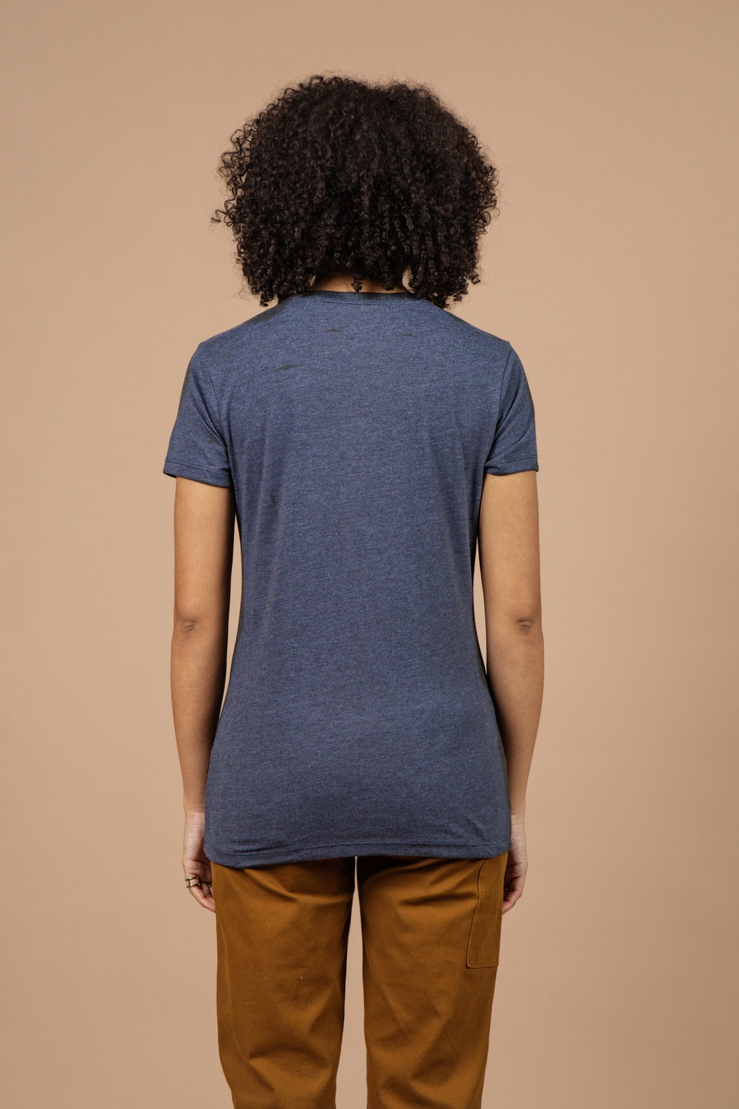 A person with curly hair, facing away from the camera, wears a Bridge & Burn Women's Peace In Plants Tee in navy and brown pants against a plain beige background.