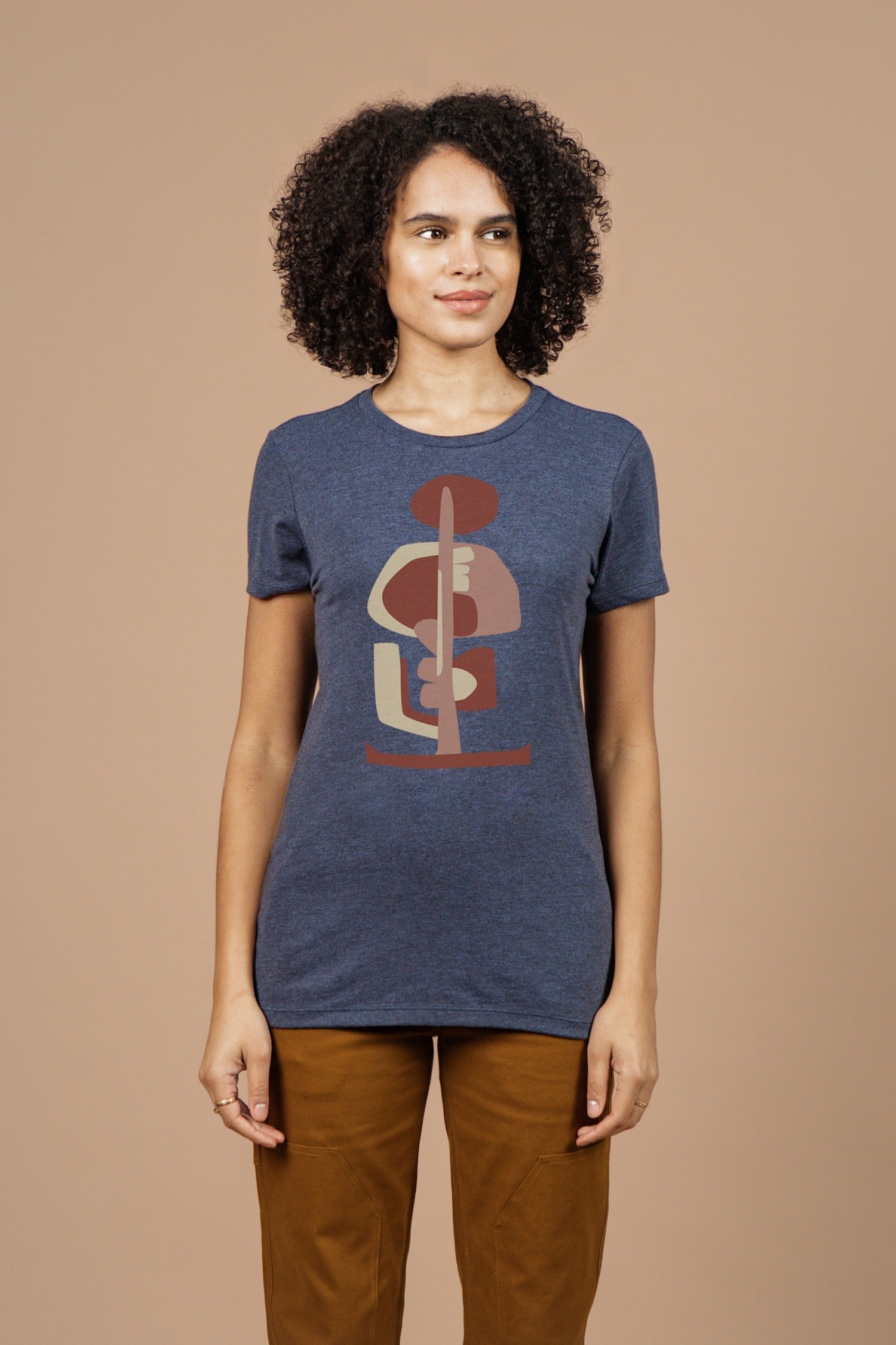 A woman with curly hair wears Bridge & Burn’s Women’s Tallest Tree Tee in navy. The eco-friendly shirt features an abstract geometric design and stands out as she smiles slightly against a plain beige background.