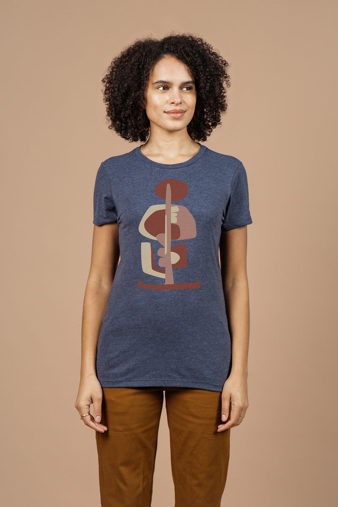 A woman with curly hair wears Bridge & Burn’s Women’s Tallest Tree Tee in navy. The eco-friendly shirt features an abstract geometric design and stands out as she smiles slightly against a plain beige background.