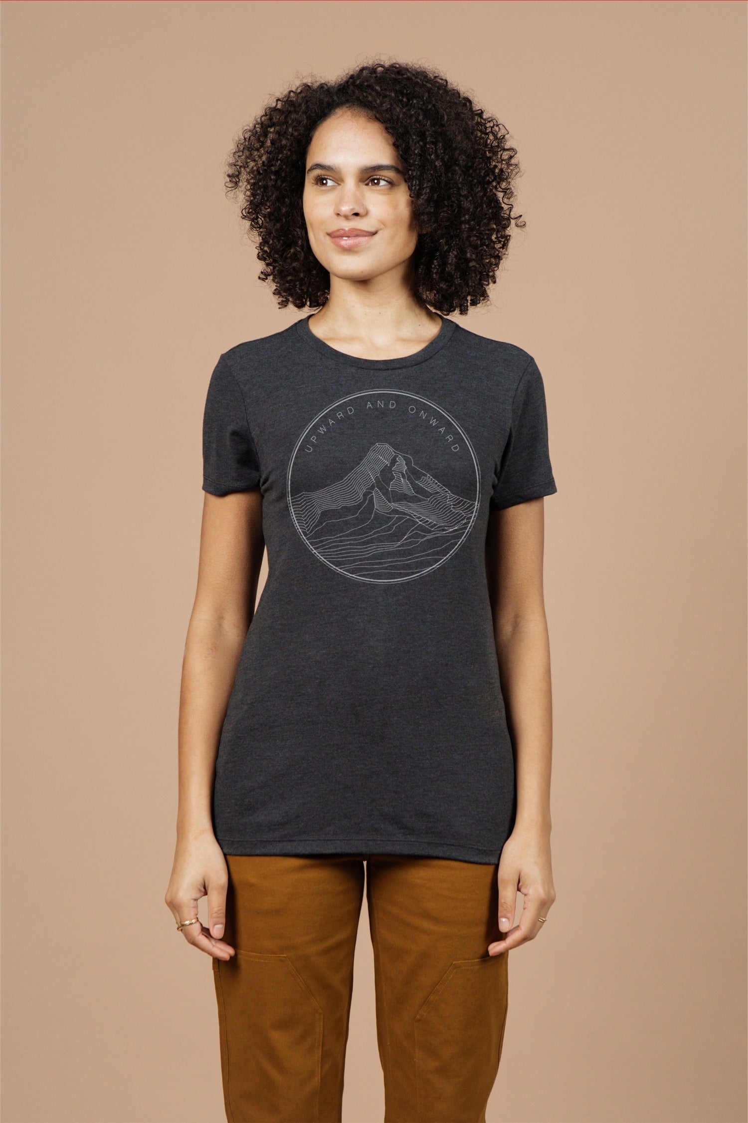 A woman with curly hair wears the Bridge & Burn Women's Upward and Onward Tee in black—an eco-friendly shirt with a minimalist mountain graphic—paired with brown pants against a plain beige background.