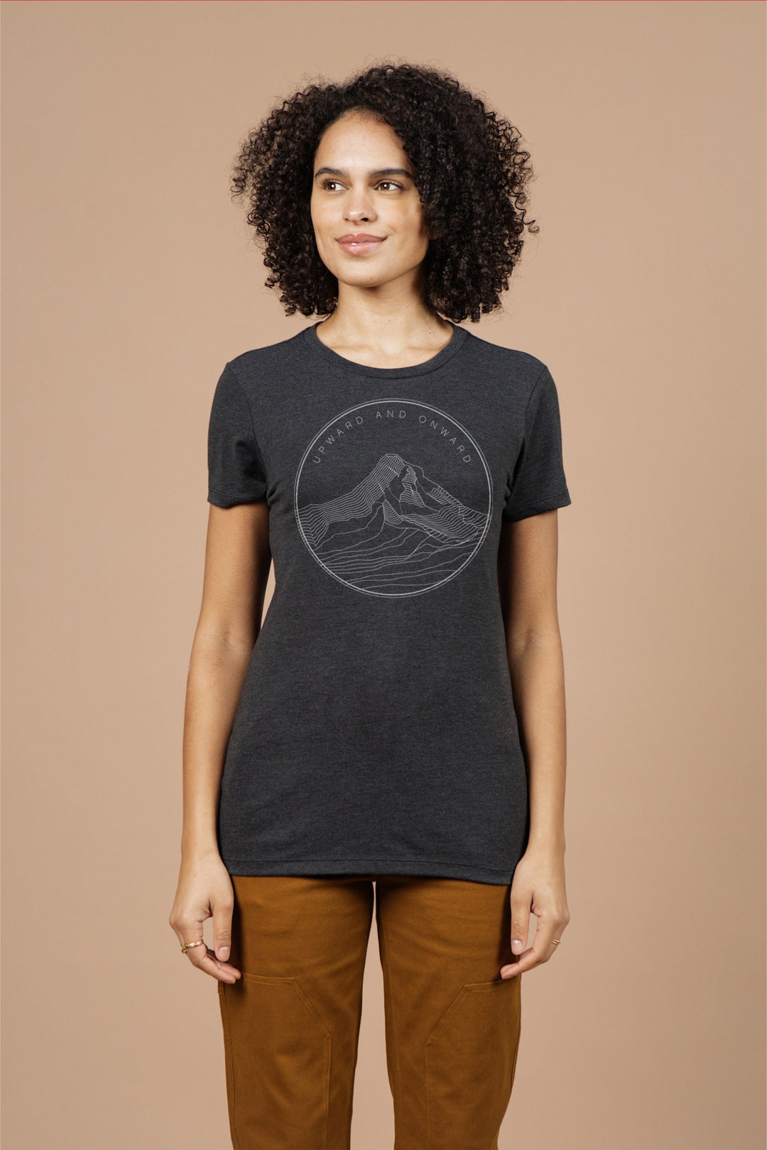 A woman with curly hair wears the Bridge & Burn Women's Upward and Onward Tee in black—an eco-friendly shirt with a minimalist mountain graphic—paired with brown pants against a plain beige background.