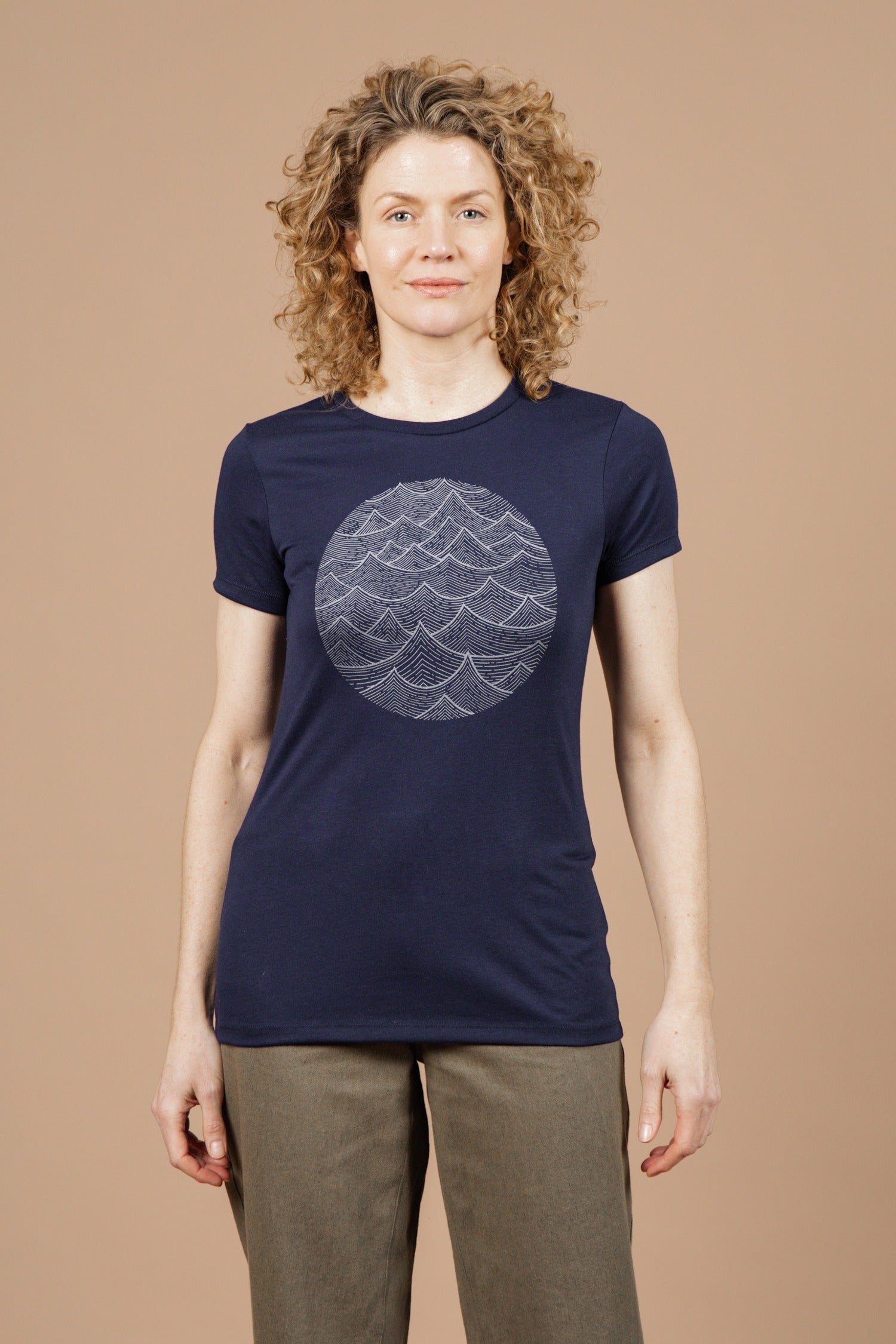 A person with curly hair wears the Bridge & Burn Women's Waves Tee in navy—made from organic cotton with a white circular wave design—paired with olive green pants, standing against a plain beige background.