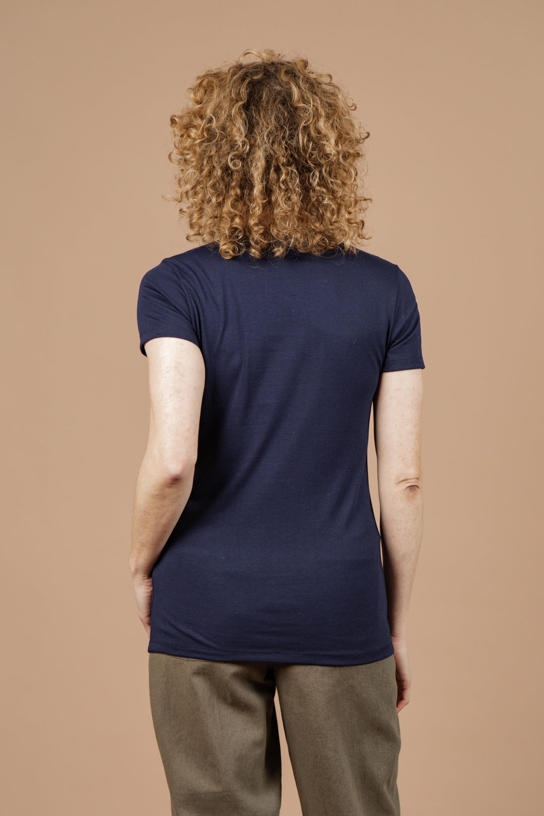 A person with curly hair faces away from the camera, wearing the Bridge & Burn Women's Waves Tee in navy and olive green pants, set against a plain beige background. The tee is lightweight and made from recycled plastic bottles.