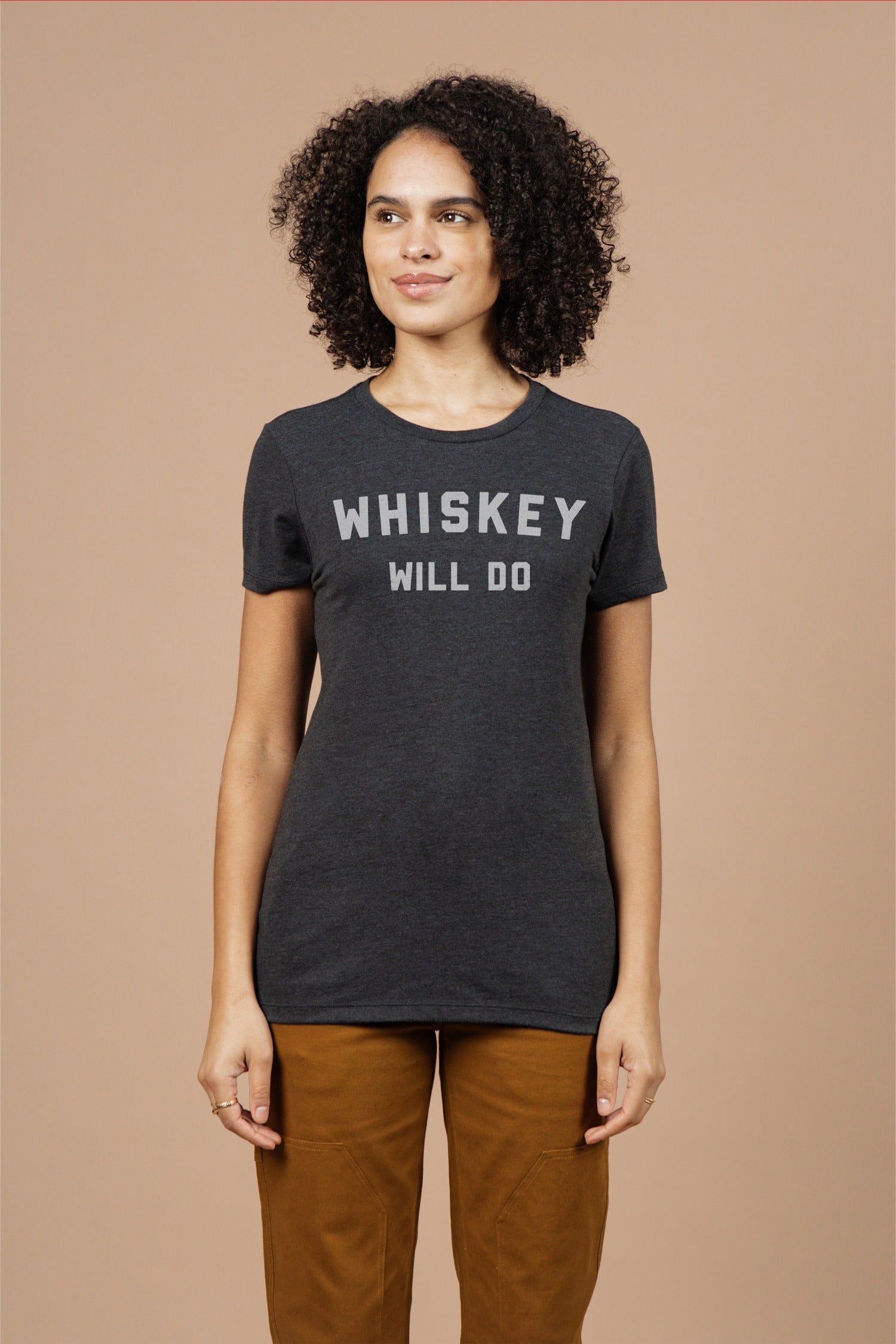 A woman with curly hair wears the Bridge & Burn Women's Whiskey Will Do Tee in black, featuring white lettering. She pairs it with brown pants and smiles softly while standing against a beige background, looking to the side.