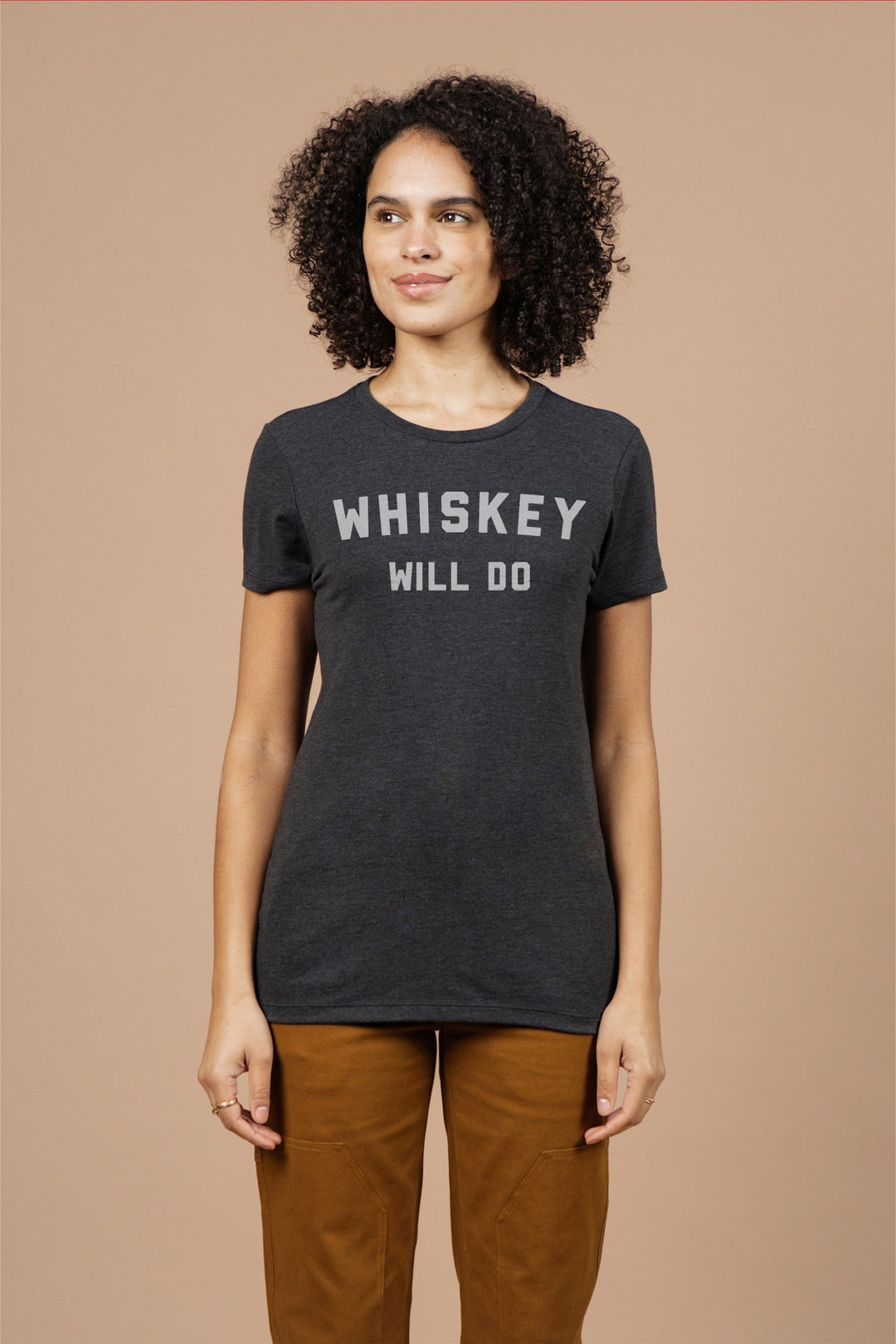 A woman with curly hair wears the Bridge & Burn Women's Whiskey Will Do Tee in black, featuring white lettering. She pairs it with brown pants and smiles softly while standing against a beige background, looking to the side.