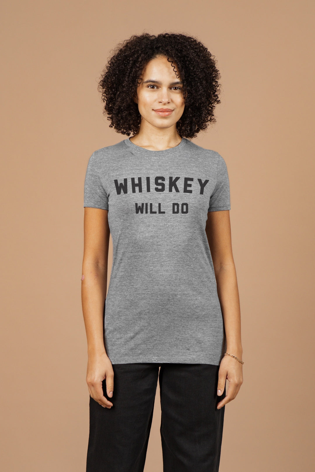 A person with curly hair wears the Bridge & Burn Women's Whiskey Will Do Tee in grey, standing against a beige background and looking at the camera with a neutral expression.