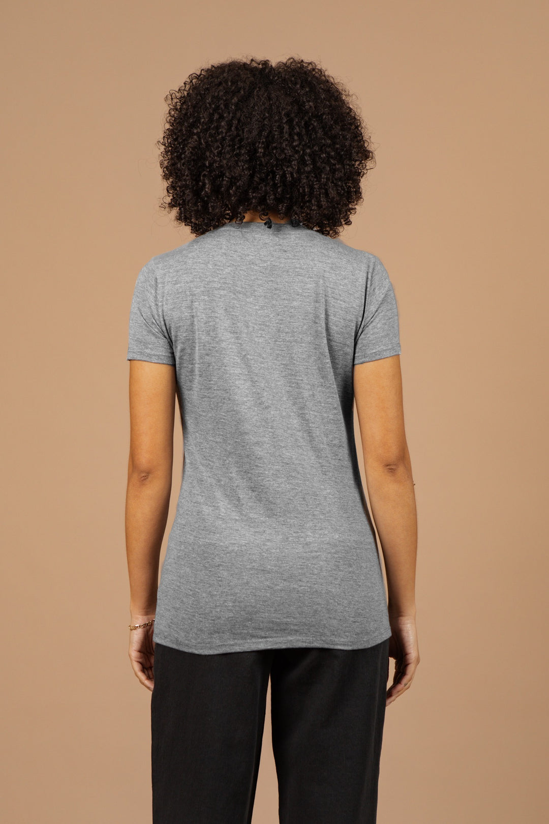 A person with curly hair is facing away from the camera, wearing the Bridge & Burn Women's Whiskey Will Do Tee in grey and black pants, against a plain beige background.