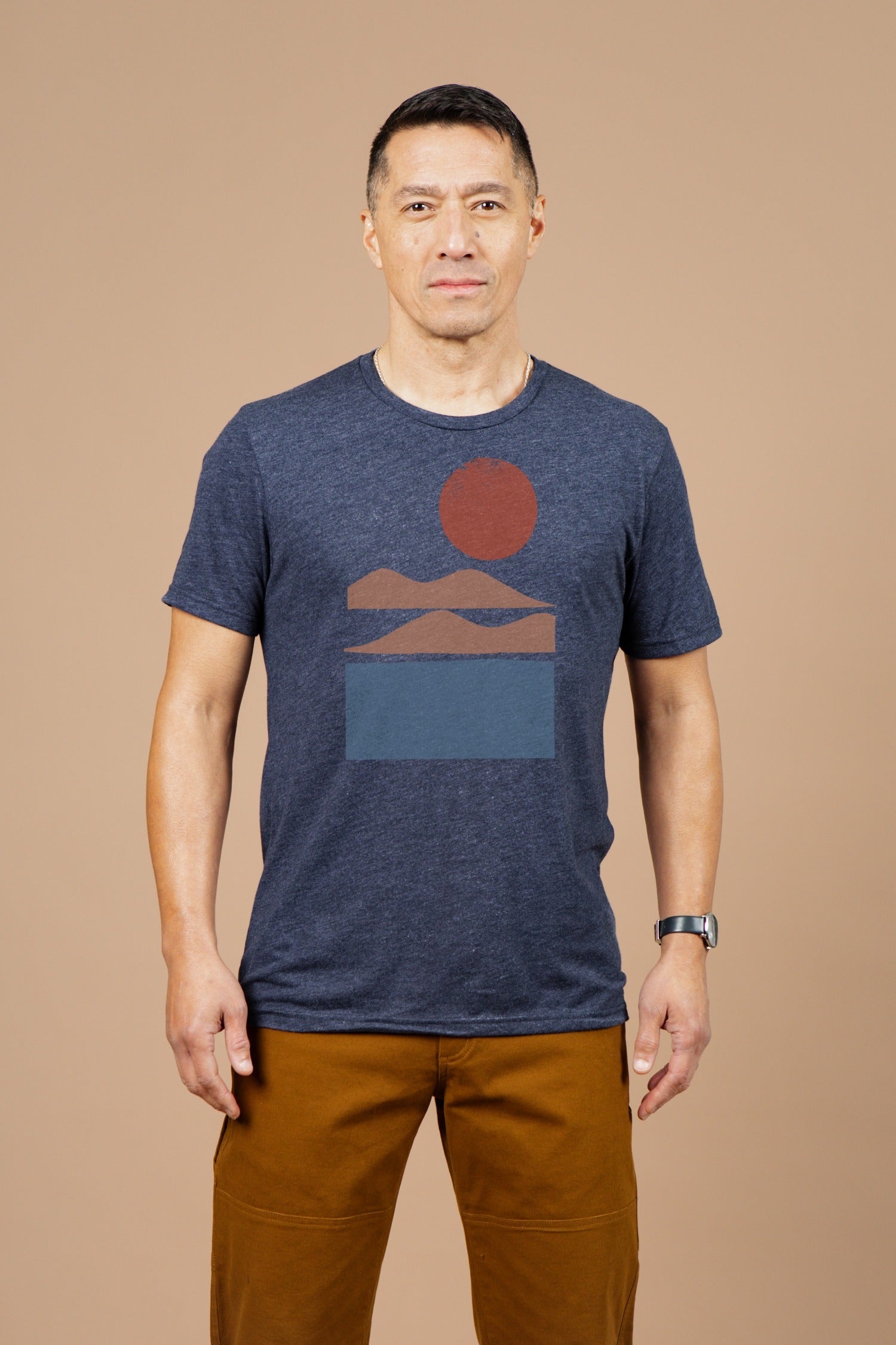 A man with short dark hair and a neutral expression wears the Bridge & Burn Men's Alpenglow Tee in navy and brown pants, standing against a plain beige background. He also has a watch on his left wrist.
