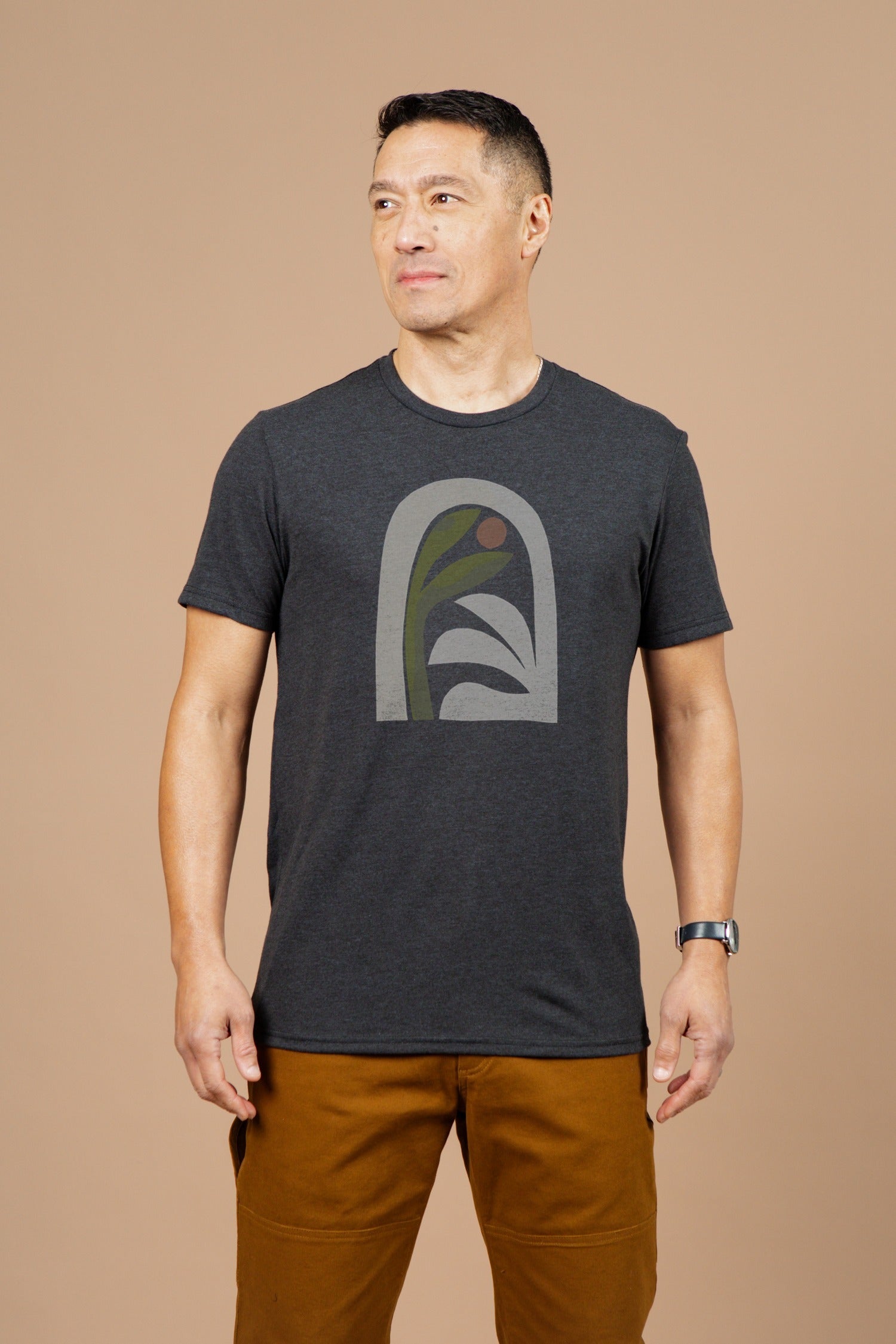 A man wearing the Bridge & Burn Men's Arch Tee in charcoal and brown pants stands against a plain beige background, looking slightly to his left.