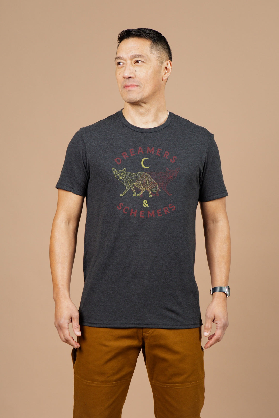 A man stands confidently against a beige background wearing the Bridge & Burn Men's Dreamers & Schemers Tee in black, which features a fox, crescent moon, and bold red and yellow lettering. He pairs it with brown pants and a watch.