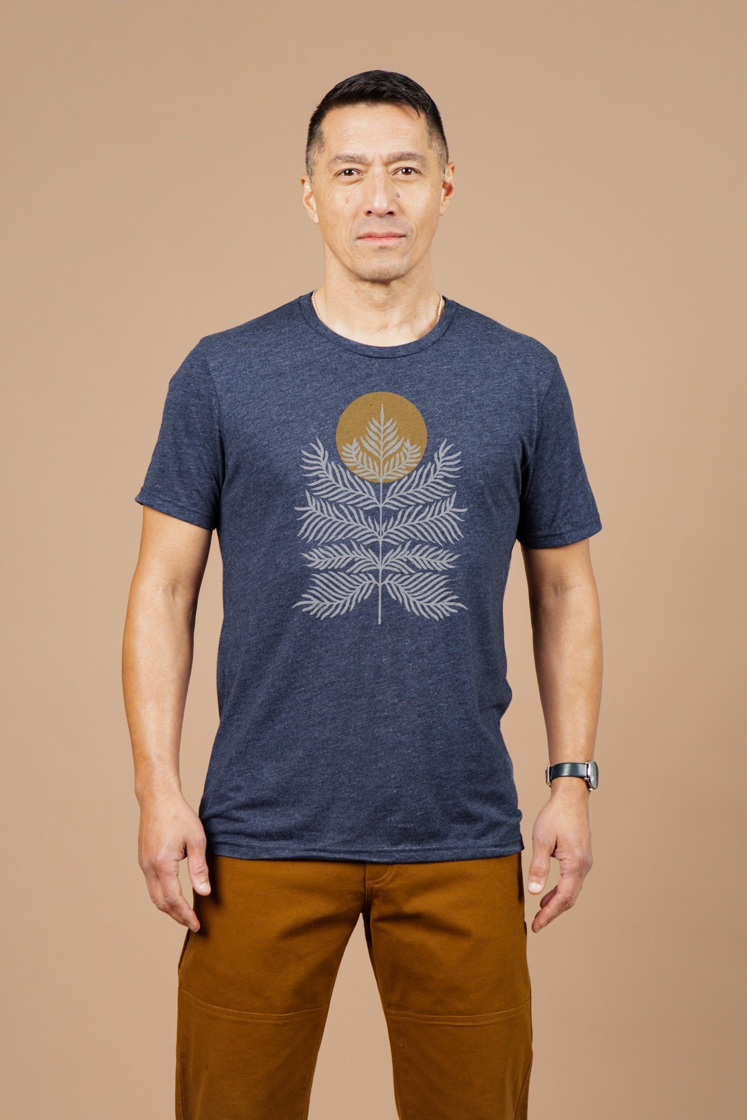 A man with short dark hair and a neutral expression stands against a beige background, wearing the Bridge & Burn Men's Peace in Plants Tee in Navy, featuring a gold circle and white fern design, paired with brown pants.