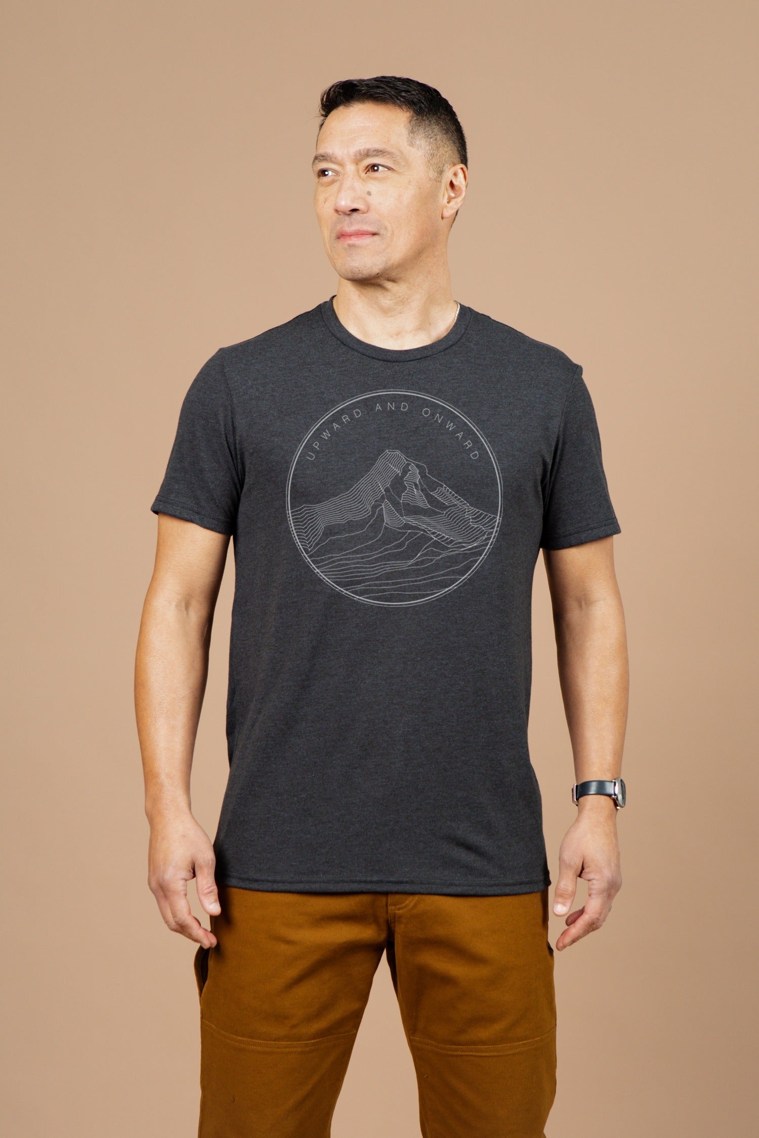 A man stands against a plain beige background, wearing the Bridge & Burn Men's Upward and Onward Tee in black, featuring a mountain graphic and slogan, paired with brown pants and a watch. He looks slightly to his left.