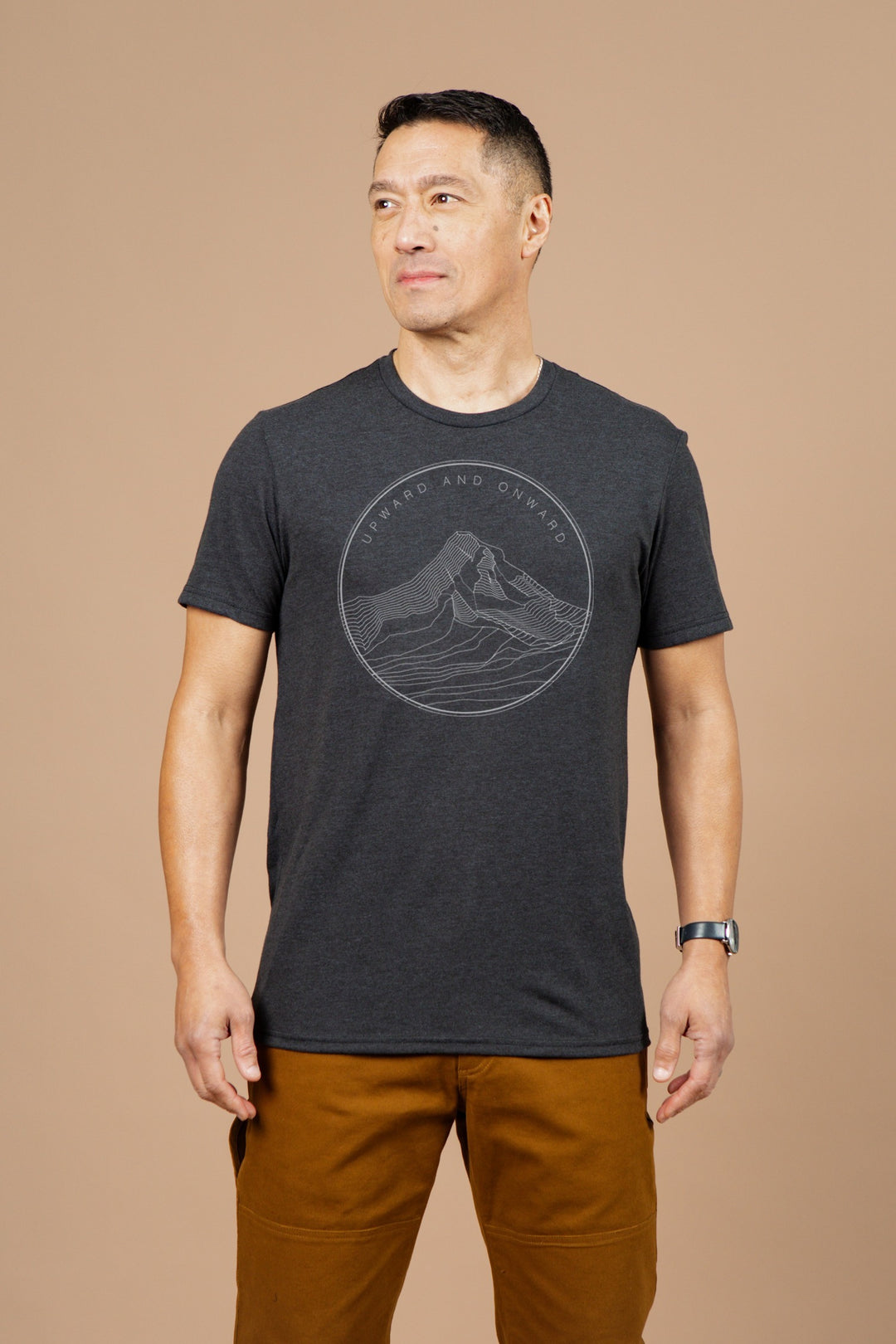 A man stands against a plain beige background, wearing the Bridge & Burn Men's Upward and Onward Tee in black, featuring a mountain graphic and slogan, paired with brown pants and a watch. He looks slightly to his left.
