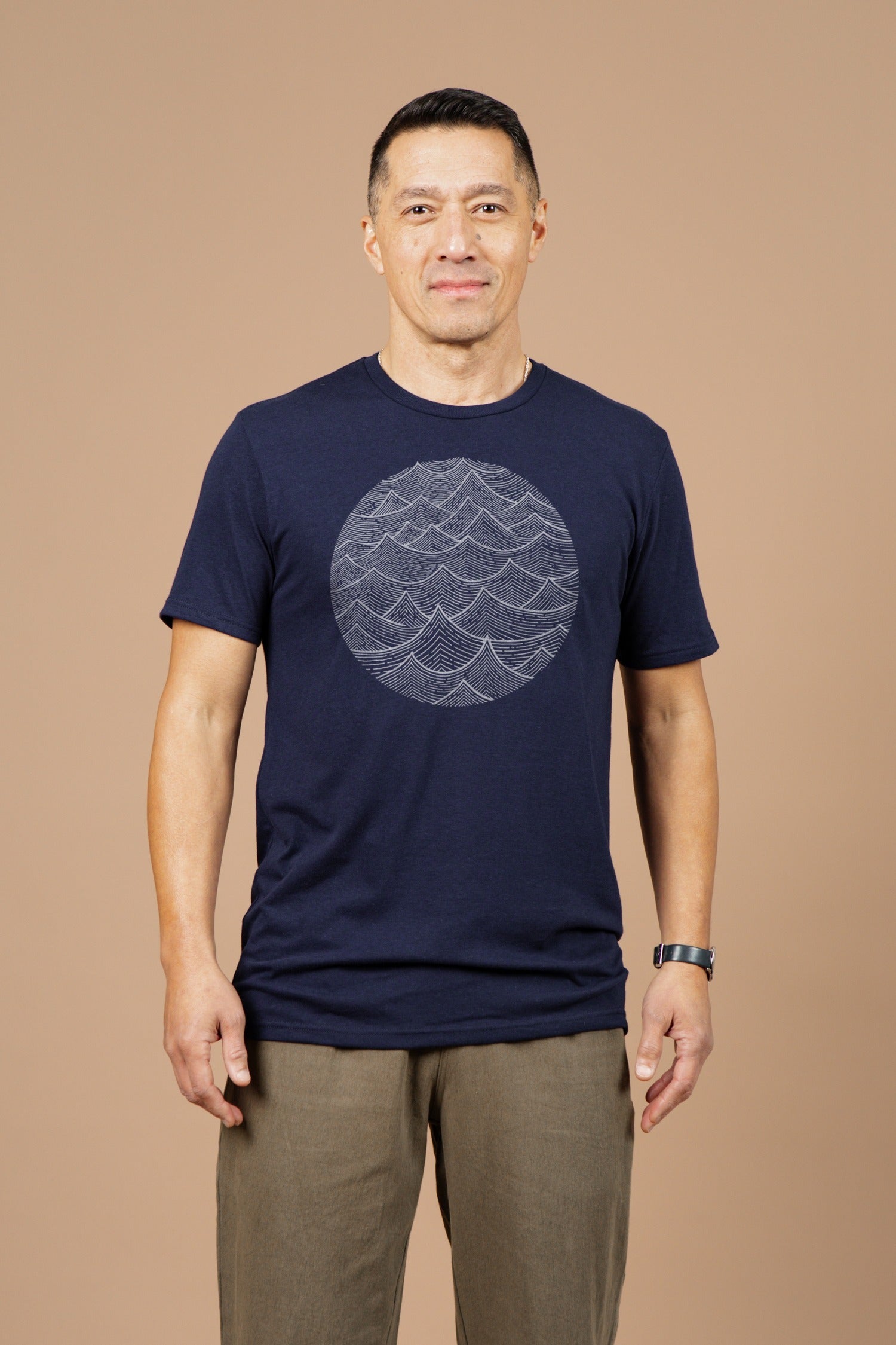 A man with short dark hair and a slight smile stands against a beige background, wearing Bridge & Burn's Men's Waves Tee in Navy—an organic cotton t-shirt featuring a circular wave pattern—paired with olive green pants.