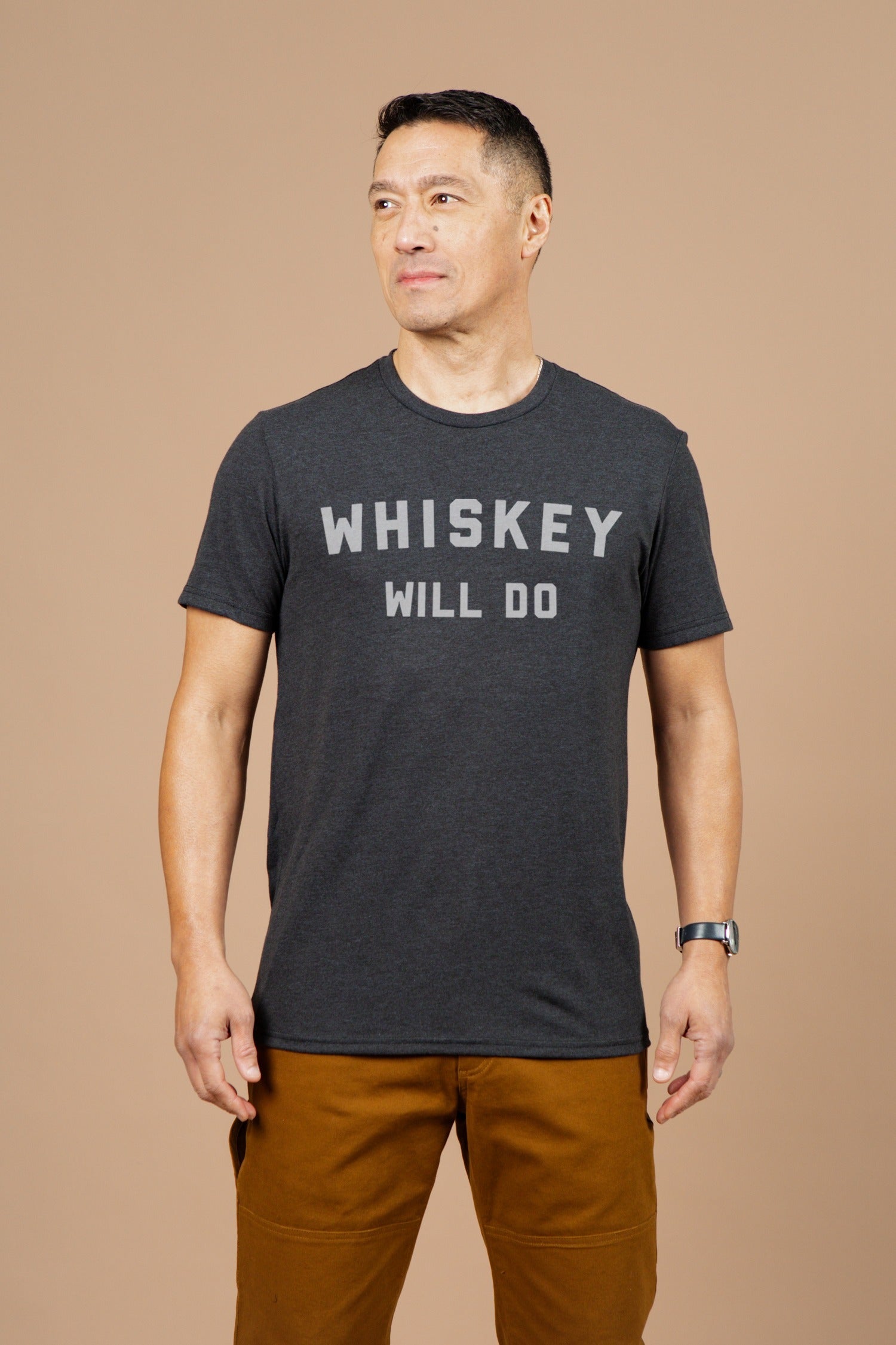 A man stands against a beige background, wearing the Bridge & Burn Men's Whiskey Will Do Tee in black with bold white lettering and brown pants. He looks slightly left and smiles softly.
