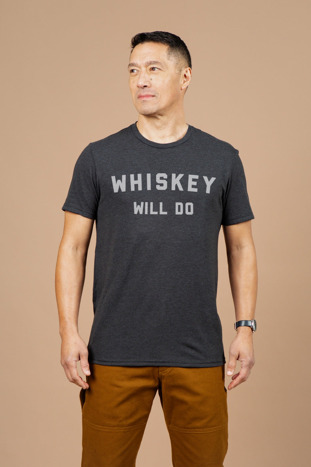 A man stands against a beige background, wearing the Bridge & Burn Men's Whiskey Will Do Tee in black with bold white lettering and brown pants. He looks slightly left and smiles softly.