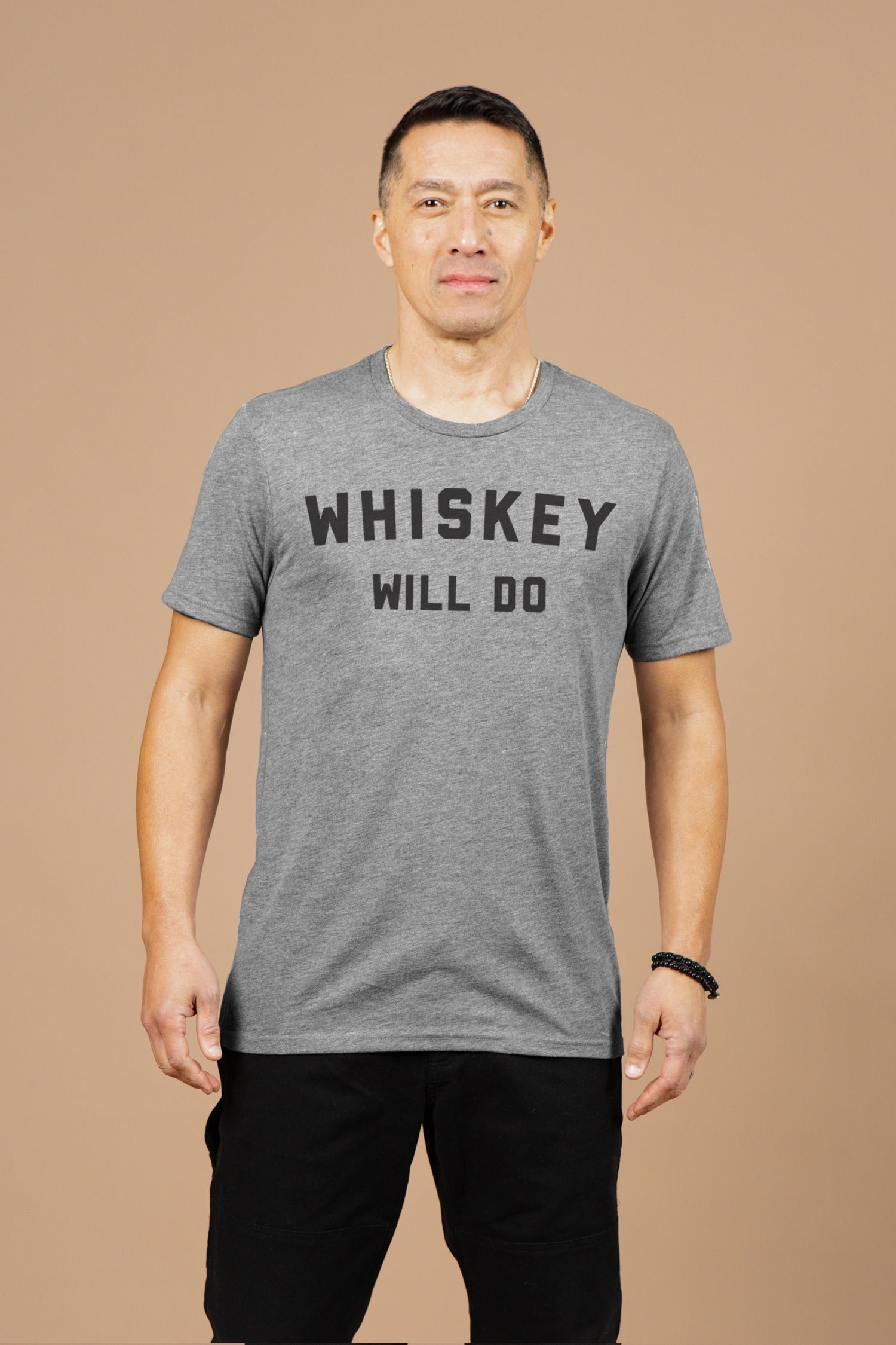 A man with short dark hair stands against a beige background, wearing the Bridge & Burn Men's Whiskey Will Do Tee in grey, featuring bold black "WHISKEY WILL DO" text, paired with black shorts.