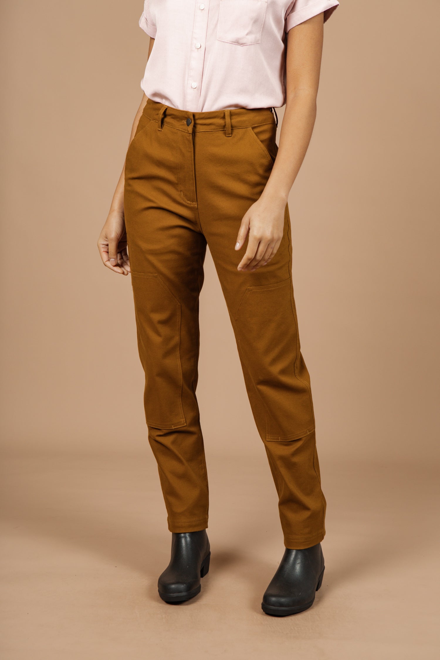A person stands against a beige background wearing high-waisted Bridge & Burn Bowden Utility Pant in Hickory Canvas with reinforced knees, a light pink short-sleeve button-up shirt, and black ankle boots. Their face is not visible.