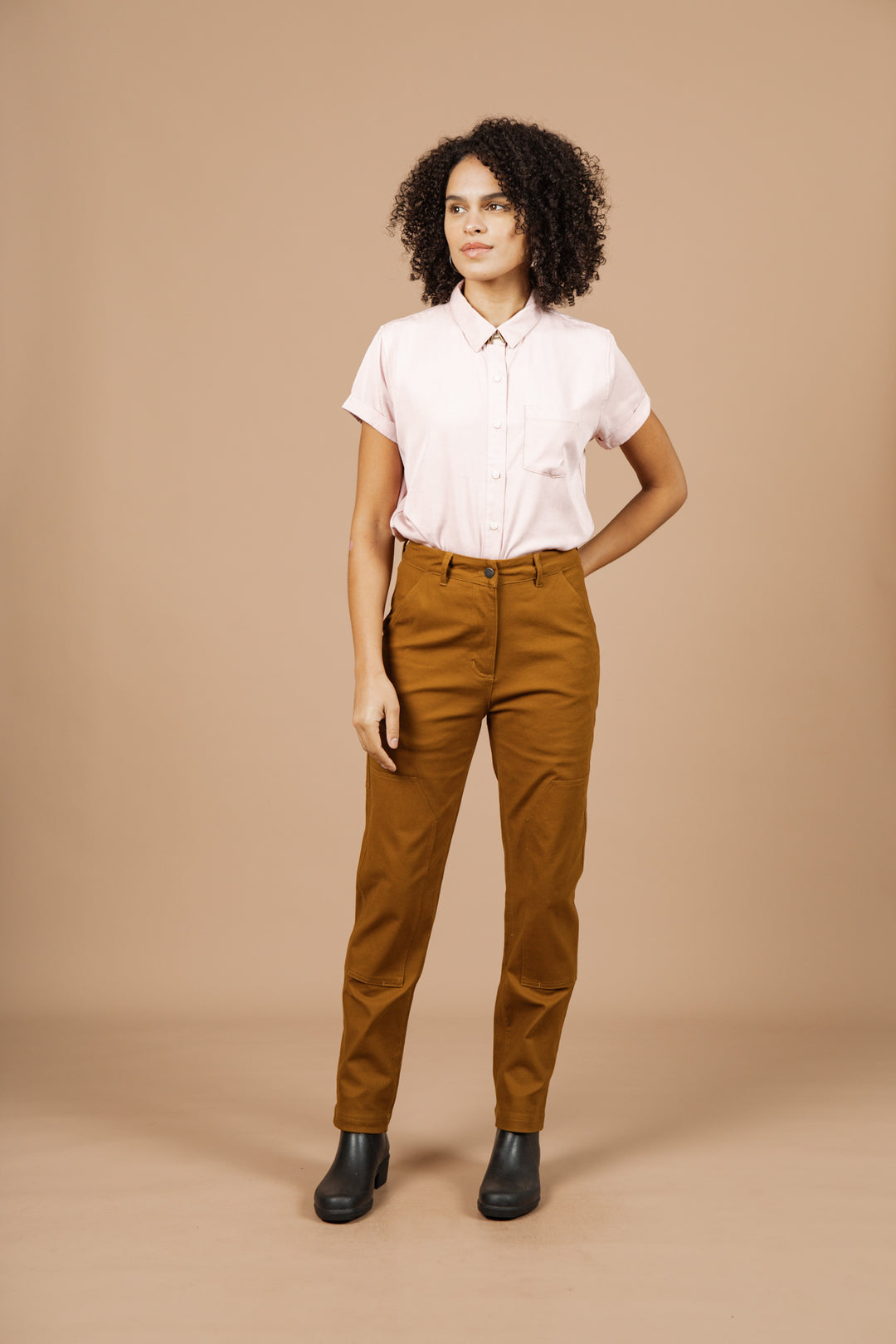 A person with curly hair stands against a beige background, wearing a light pink short-sleeved button-up shirt and Bridge & Burn Bowden Utility Pant in Hickory Canvas with reinforced knees, paired with black ankle boots and one hand in their pocket.