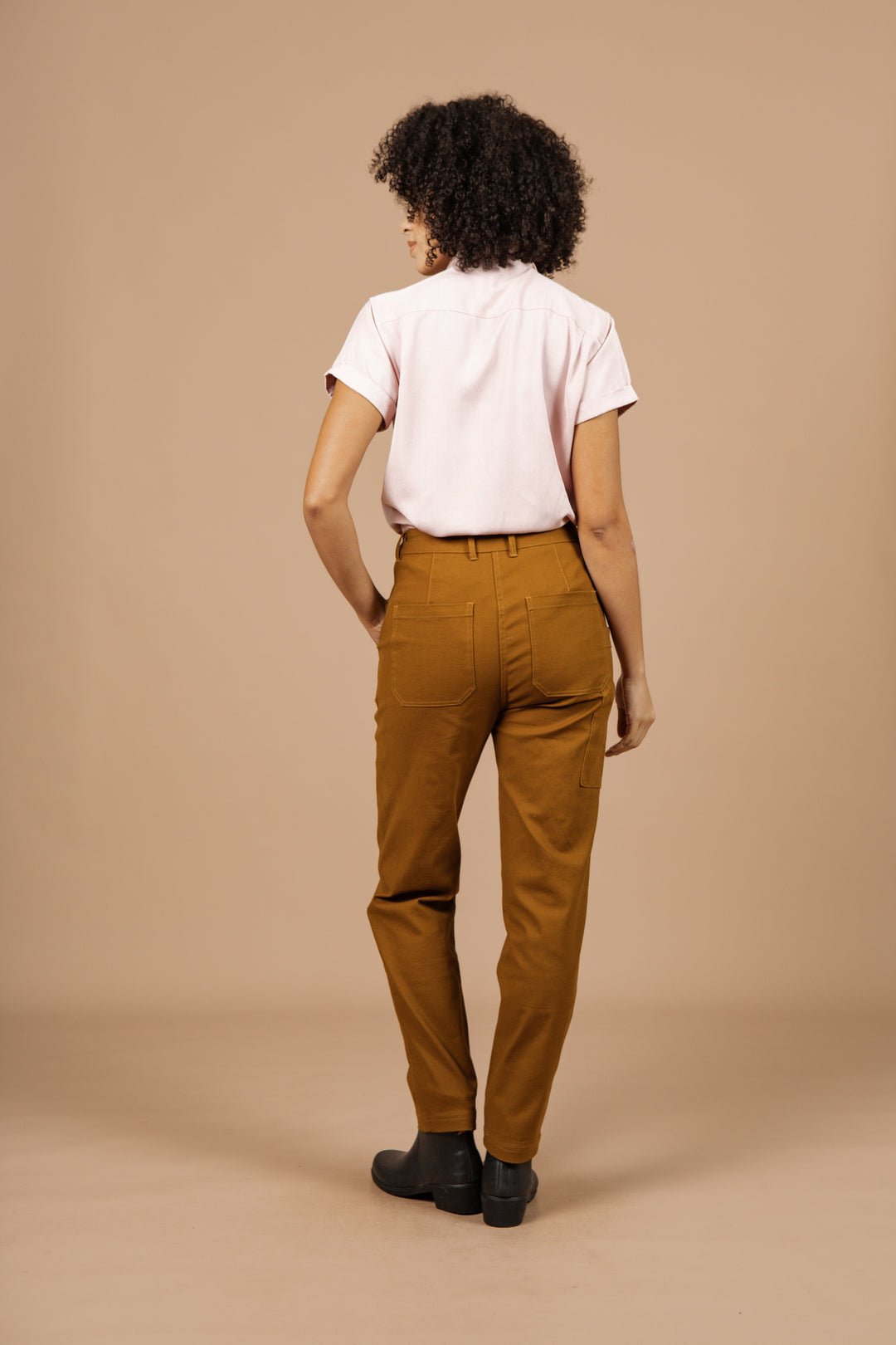 A person with curly hair, dressed in a short-sleeved light pink shirt and Bridge & Burn Bowden Utility Pant in Hickory Canvas, stands facing away from the camera against a plain beige background.