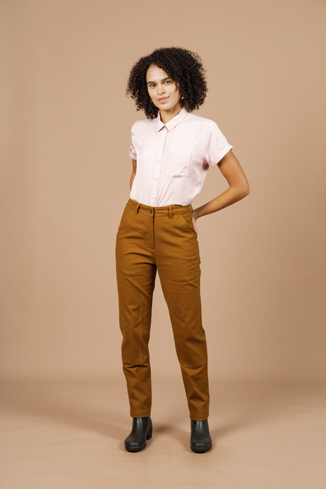 A person with curly hair stands against a beige background, wearing a light pink short-sleeved button-up shirt, Bridge & Burn’s Bowden Utility Pant in Hickory Canvas with reinforced knees, and black ankle boots, one hand behind their back.