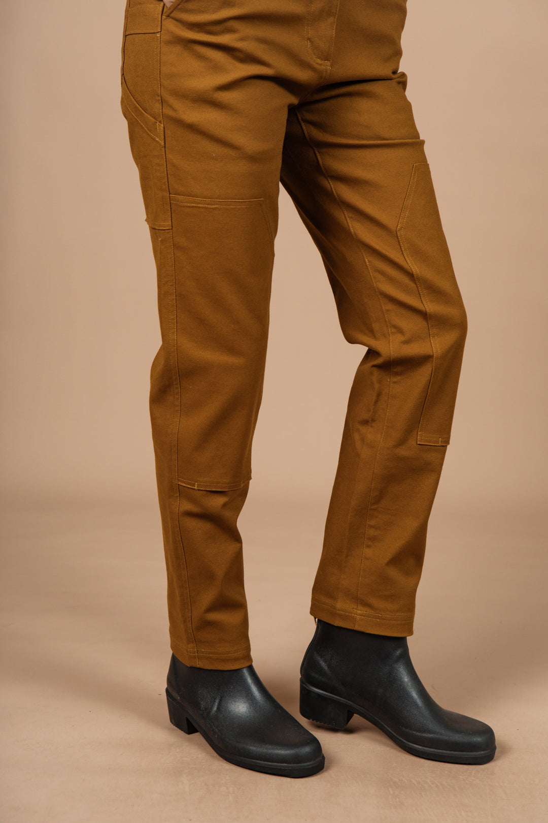 A person wears Bridge & Burn Bowden Utility Pant in Hickory Canvas with black boots, standing on a light brown surface; only their lower half is visible against a plain beige background.
