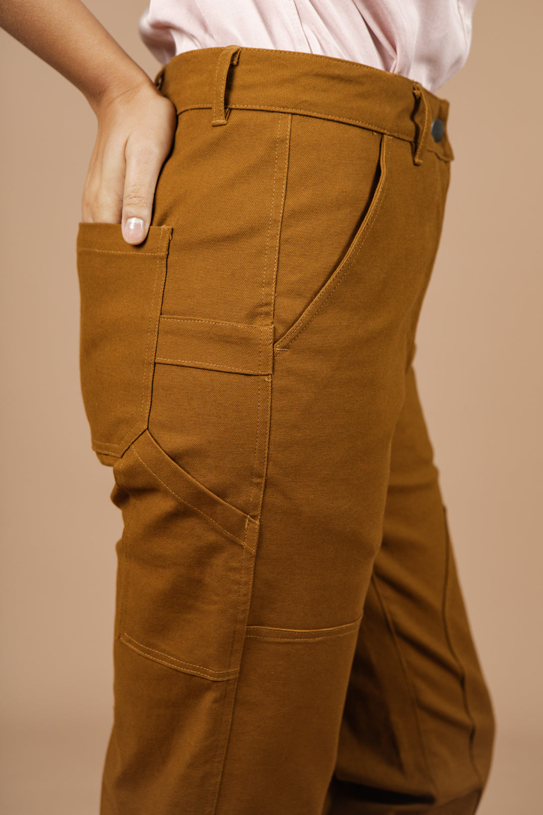 A person wears the Bridge & Burn Bowden Utility Pant in Hickory Canvas, featuring reinforced knees, paired with a light pink shirt, standing with one hand in their pocket against a plain beige background.