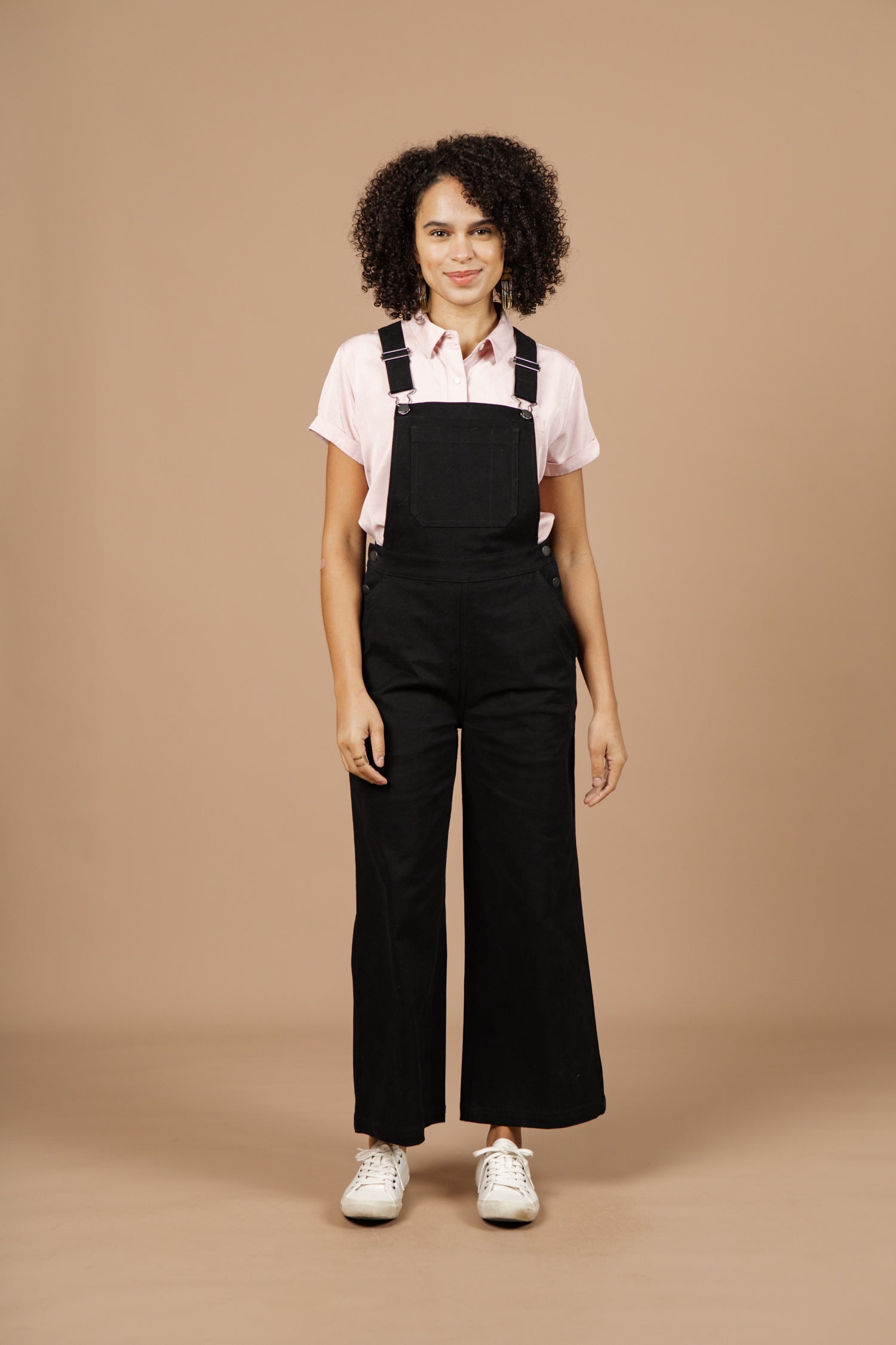 A person with curly hair smiles against a beige background, wearing a light pink short-sleeve shirt, Bridge & Burn's Roscoe Overall in Black Canvas, and white sneakers, with arms relaxed at their sides.