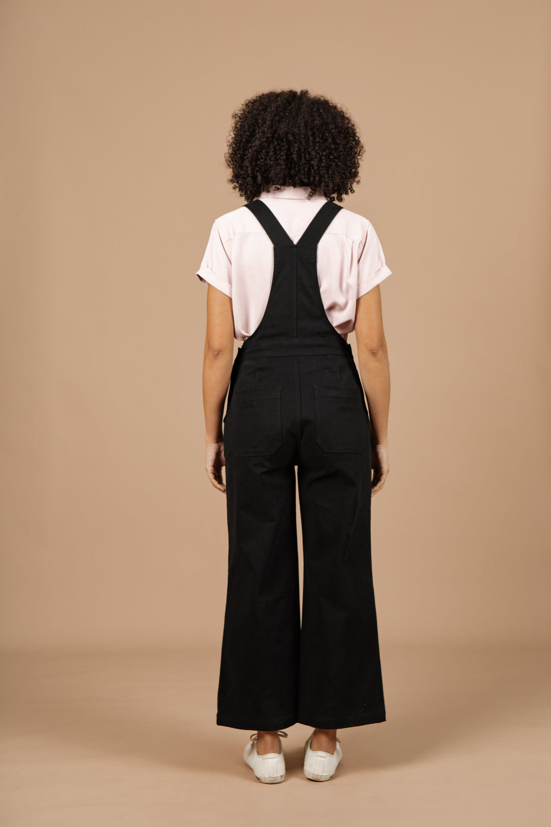 A person with curly hair, facing away, wears a light pink short-sleeve shirt and Bridge & Burn's Roscoe Overall in Black Canvas paired with white shoes, standing against a plain beige background.