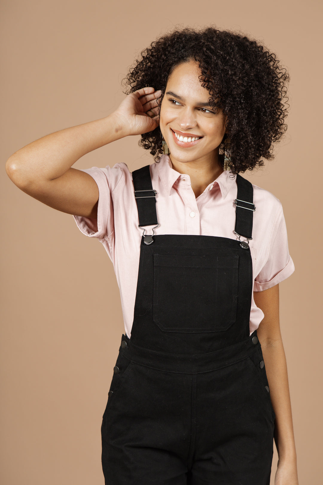 A smiling person with curly hair wears a light pink shirt and Bridge & Burn’s Roscoe Overall in black canvas, standing before a beige background with one hand touching their head.