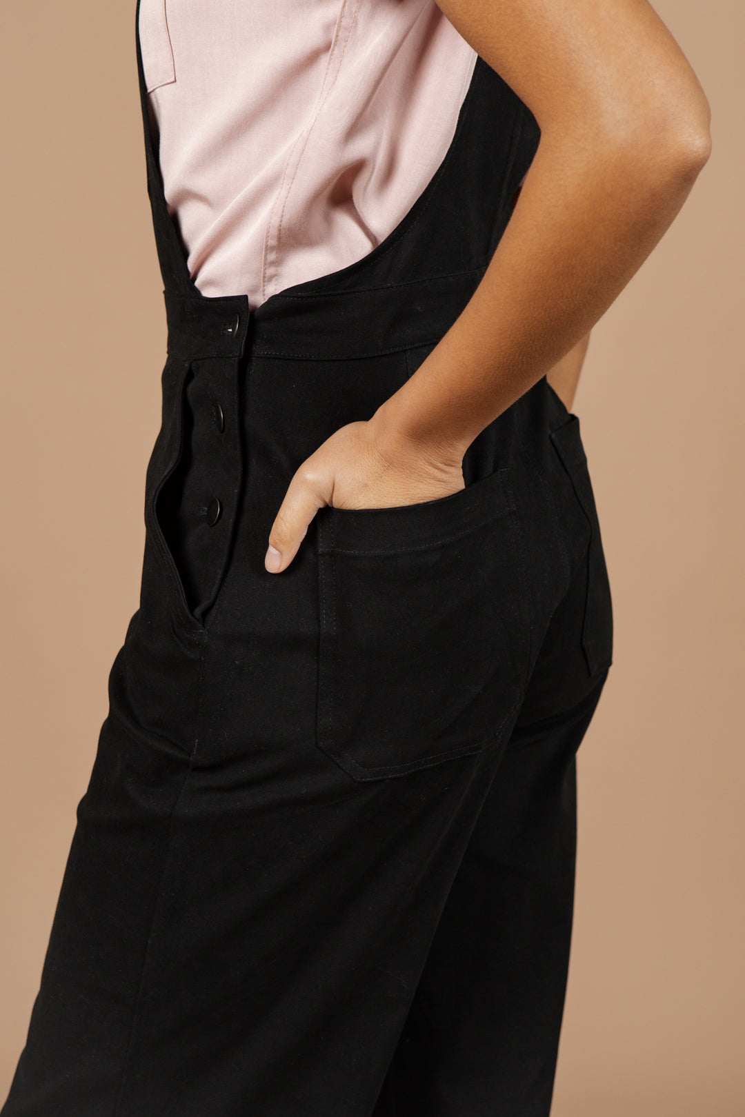 A person stands sideways with one hand in their pocket wearing Bridge & Burn's Roscoe Overall in Black Canvas and a light pink shirt, set against a plain beige background.