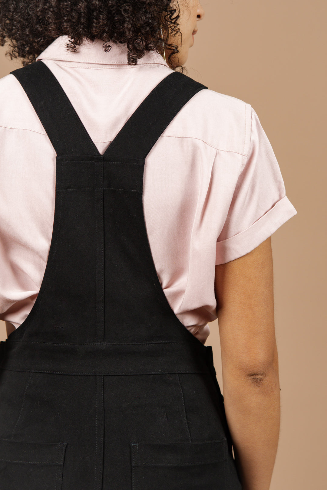 A person with curly hair, wearing Bridge & Burn’s Roscoe Overall in Black Canvas and a light pink short-sleeve shirt, is shown from the back against a beige background.