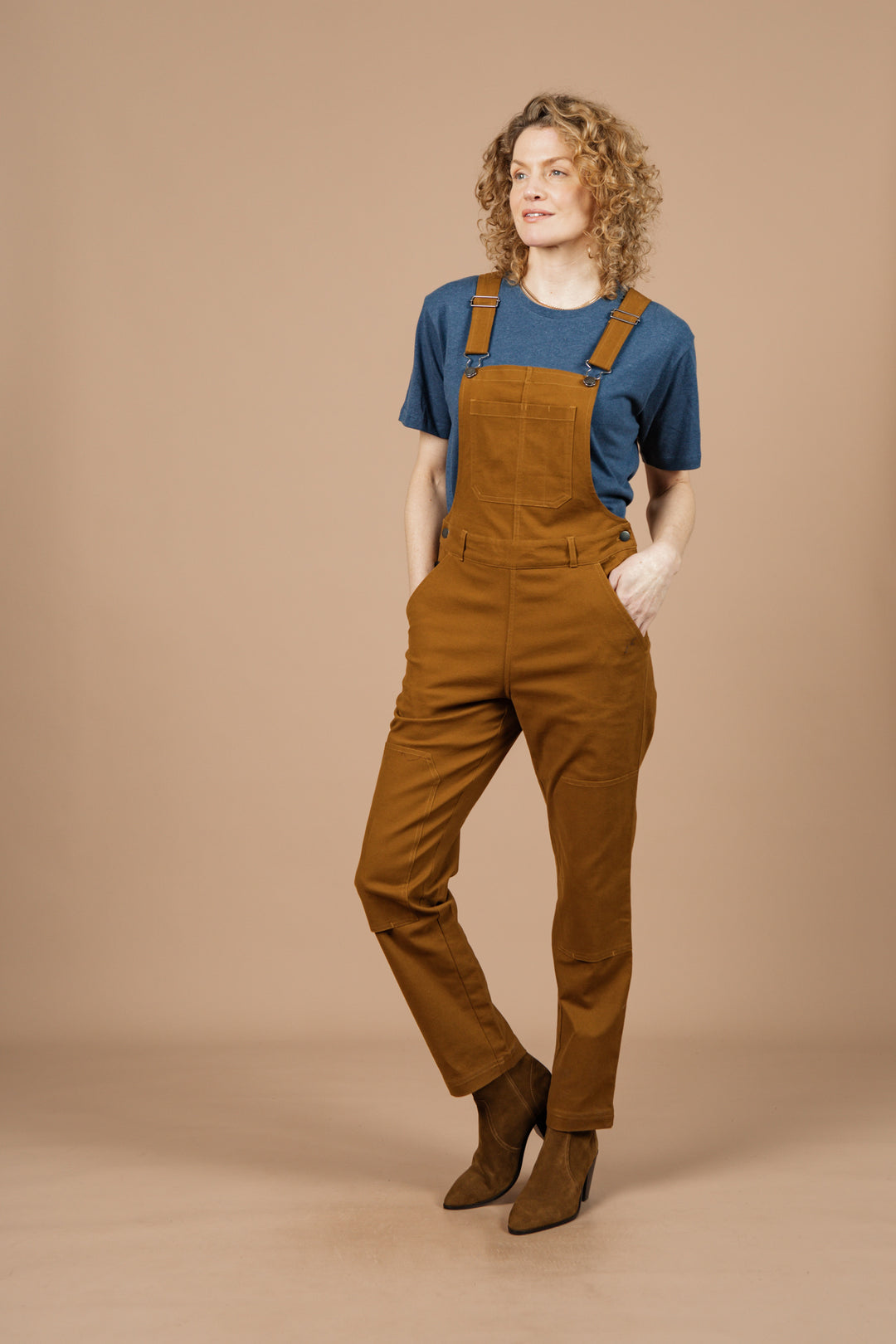 A person with curly blonde hair stands against a beige background, wearing Bridge & Burn's Tobin Utility Overall in brown canvas over a blue t-shirt, paired with brown boots, hands in pockets and a slight smile on their face.