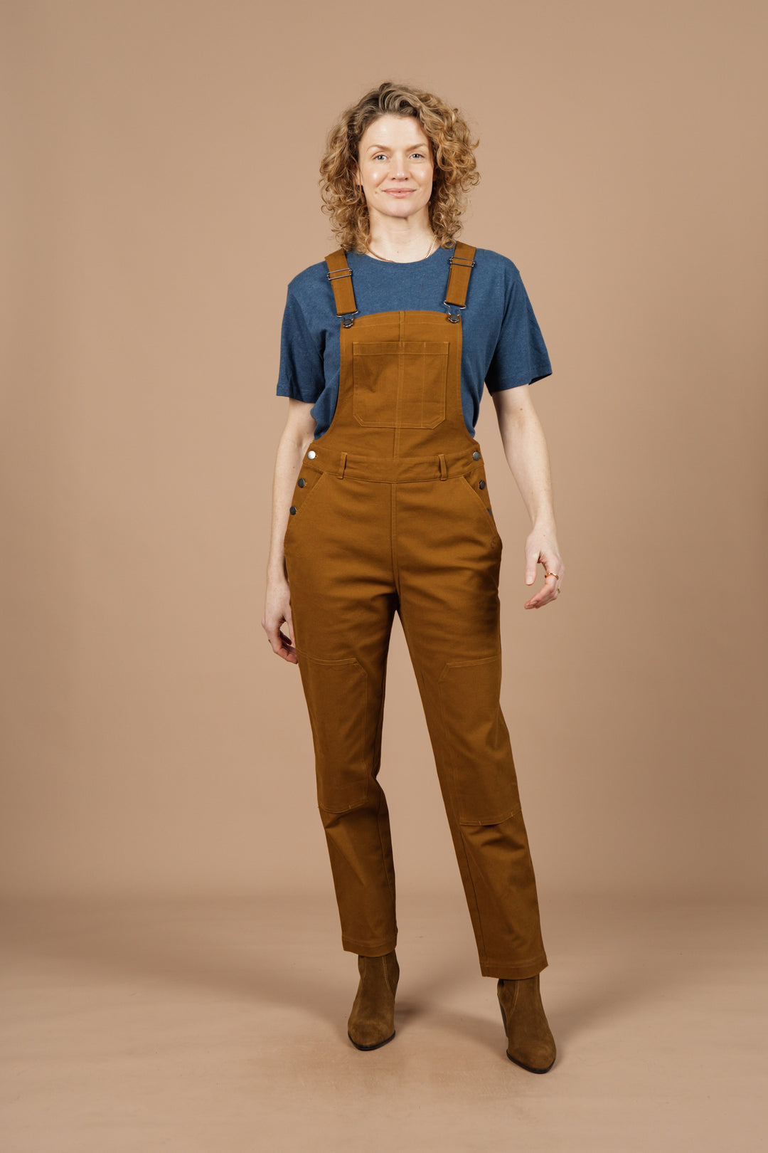 A person with curly blonde hair, wearing a blue t-shirt and Bridge & Burn’s Tobin Utility Overall in brown canvas, stands facing forward against a plain beige background.