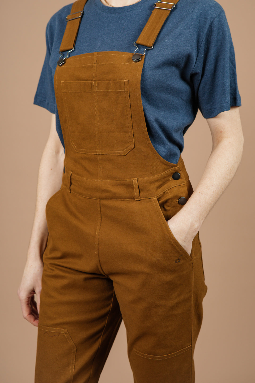 A person wearing a blue short-sleeve t-shirt and Bridge & Burn's Tobin Utility Overall in brown canvas stands with one hand in the pocket against a plain beige background. Their head is not visible.