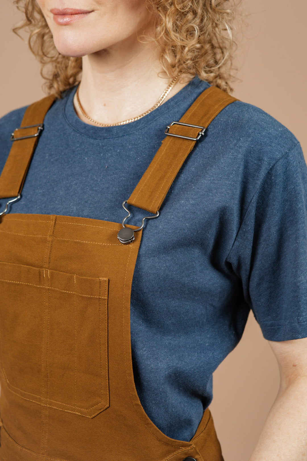 A person with curly blond hair wears a blue t-shirt and Bridge & Burn Tobin Utility Overall in Brown Canvas, standing against a beige background and facing slightly left. The image captures their upper body and part of their face.