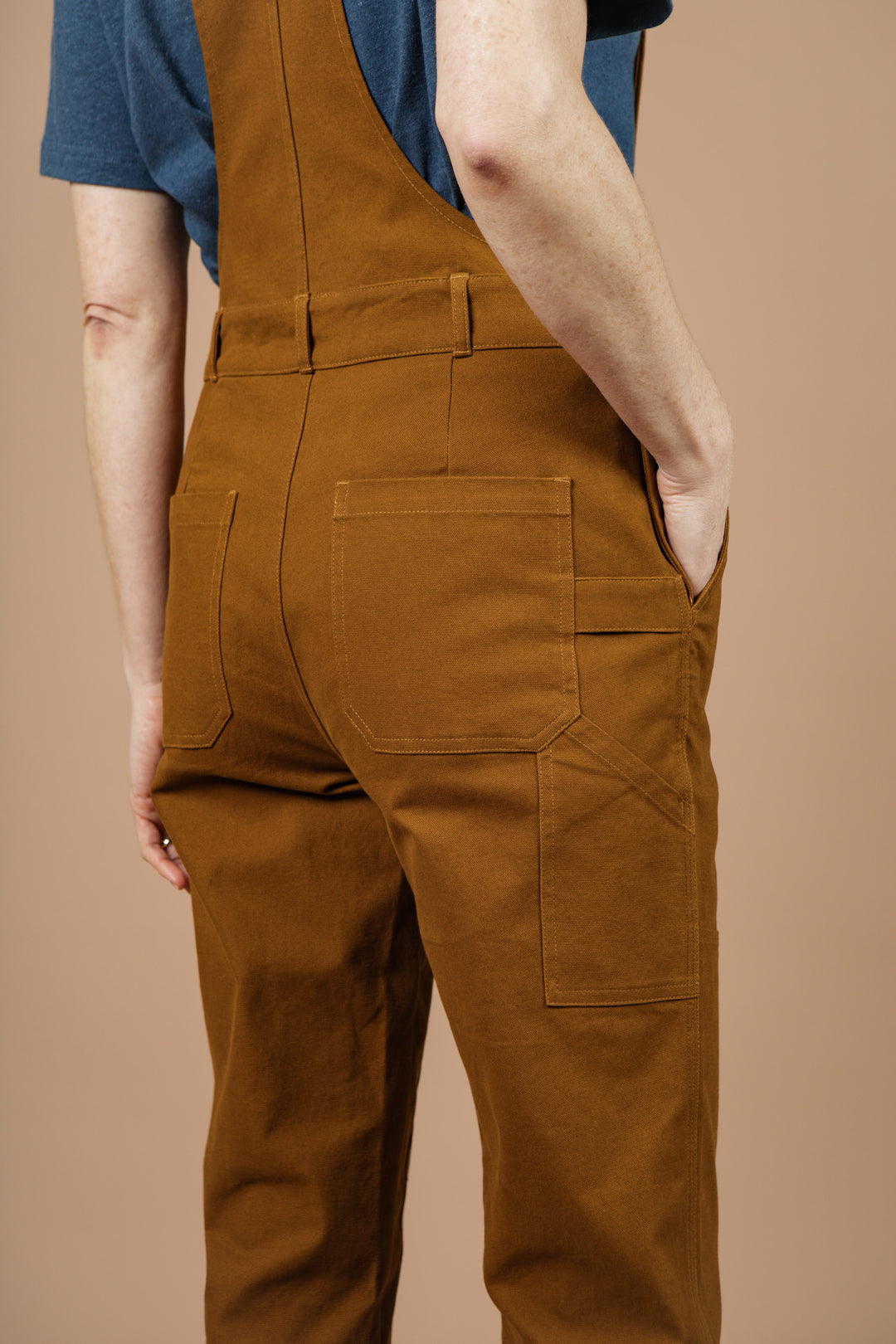 Someone stands with one hand in their pocket, shown from the back against a plain beige background, wearing the Bridge & Burn Tobin Utility Overall in brown canvas.