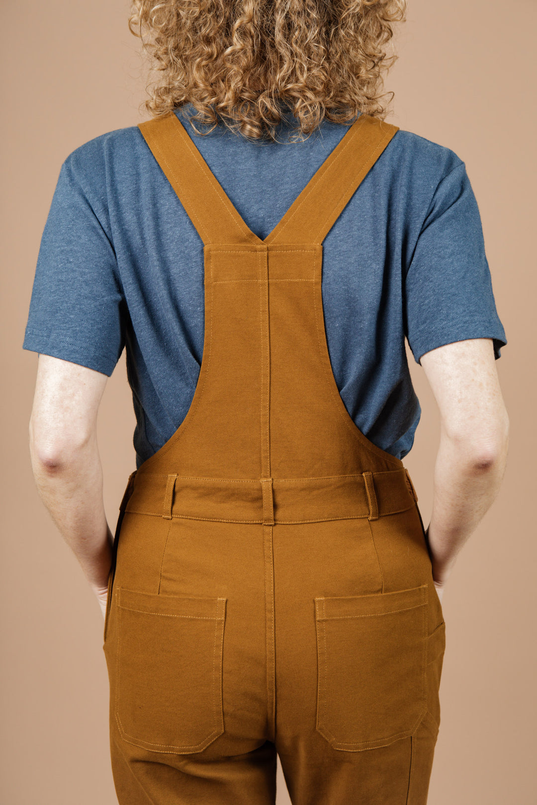 A person with curly hair, seen from behind, wears a blue t-shirt and Bridge & Burn’s Tobin Utility Overall in brown canvas. The background is solid beige.