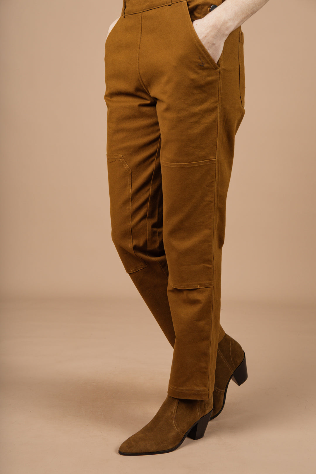 A person wears the Bridge & Burn Tobin Utility Overall in brown canvas—high-waisted with visible front seams and pockets—paired with brown suede heeled boots, standing against a beige background.