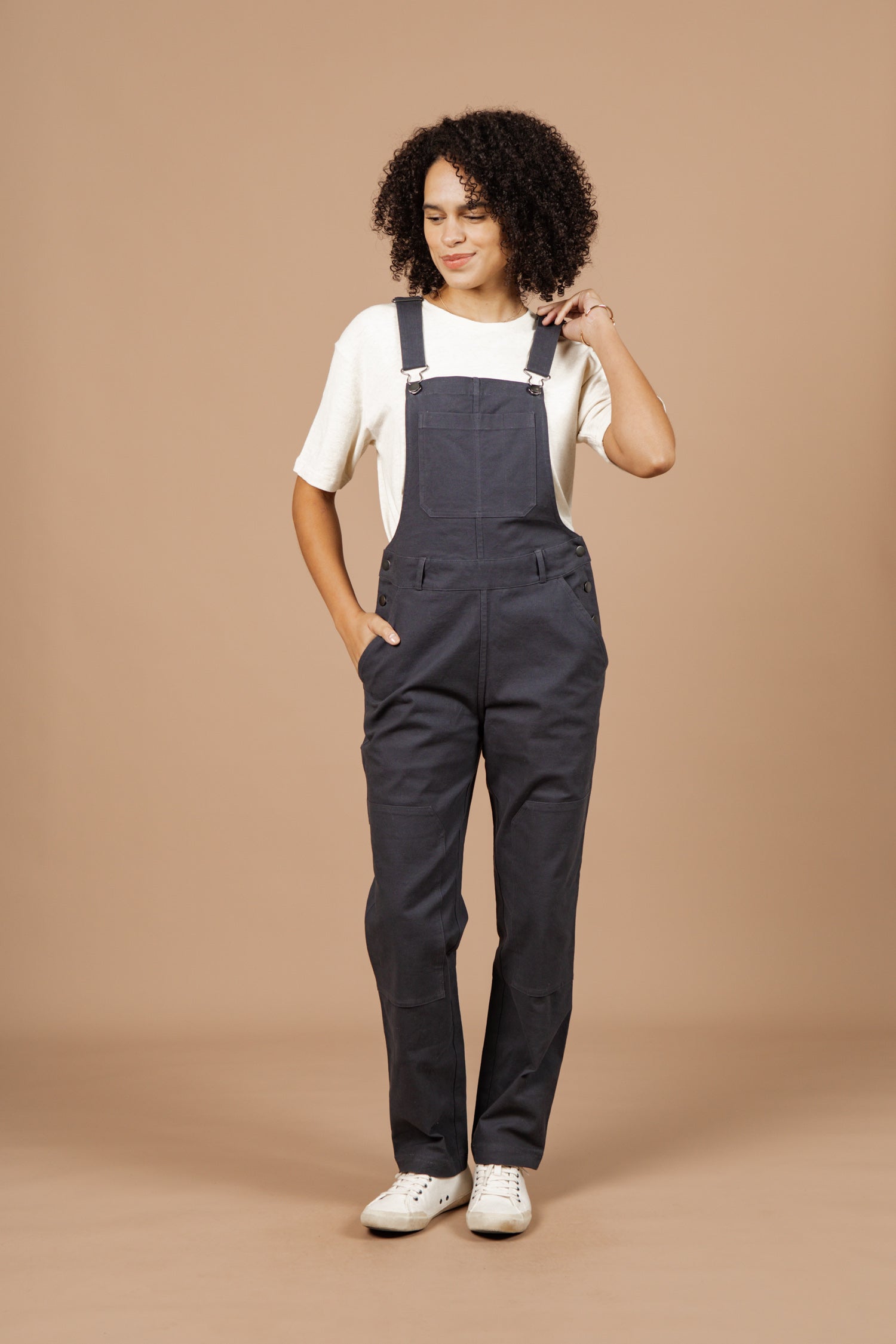 A person with curly hair wears Bridge & Burn's Tobin Utility Overall in dark charcoal canvas over a white short-sleeve shirt and white sneakers, posing against a plain beige background with one hand in their pocket and the other touching their hair.