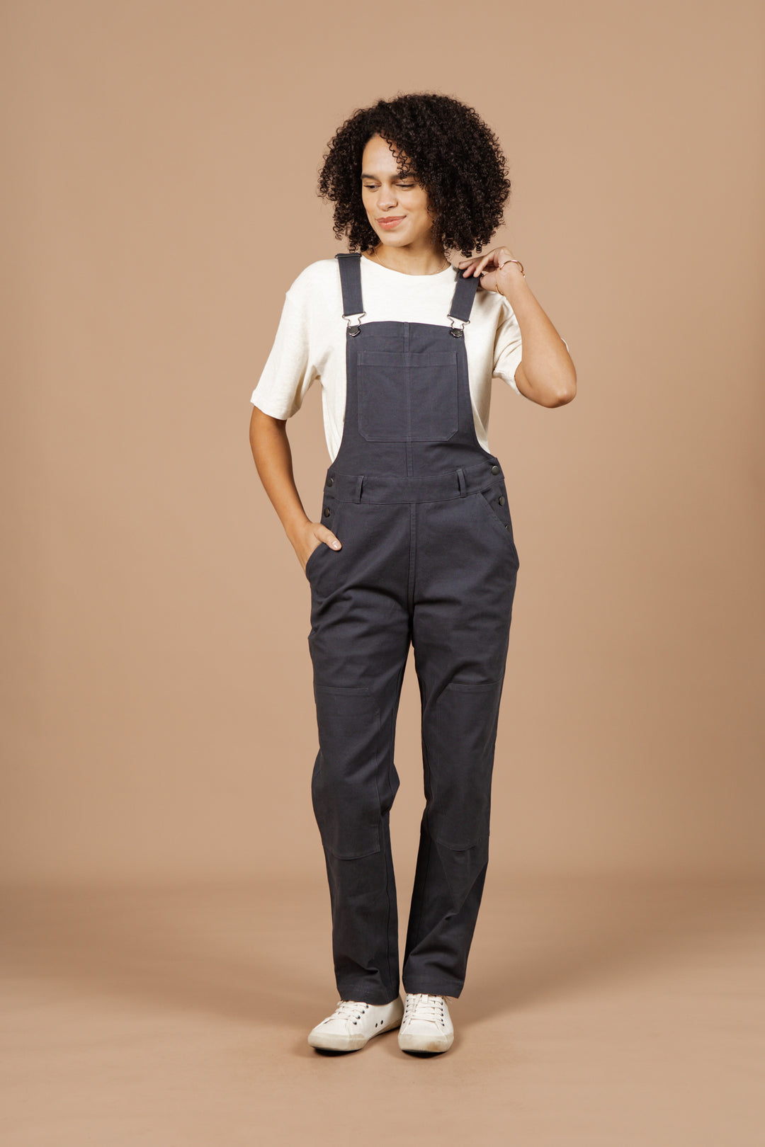 A person with curly hair wears Bridge & Burn's Tobin Utility Overall in dark charcoal canvas over a white short-sleeve shirt and white sneakers, posing against a plain beige background with one hand in their pocket and the other touching their hair.