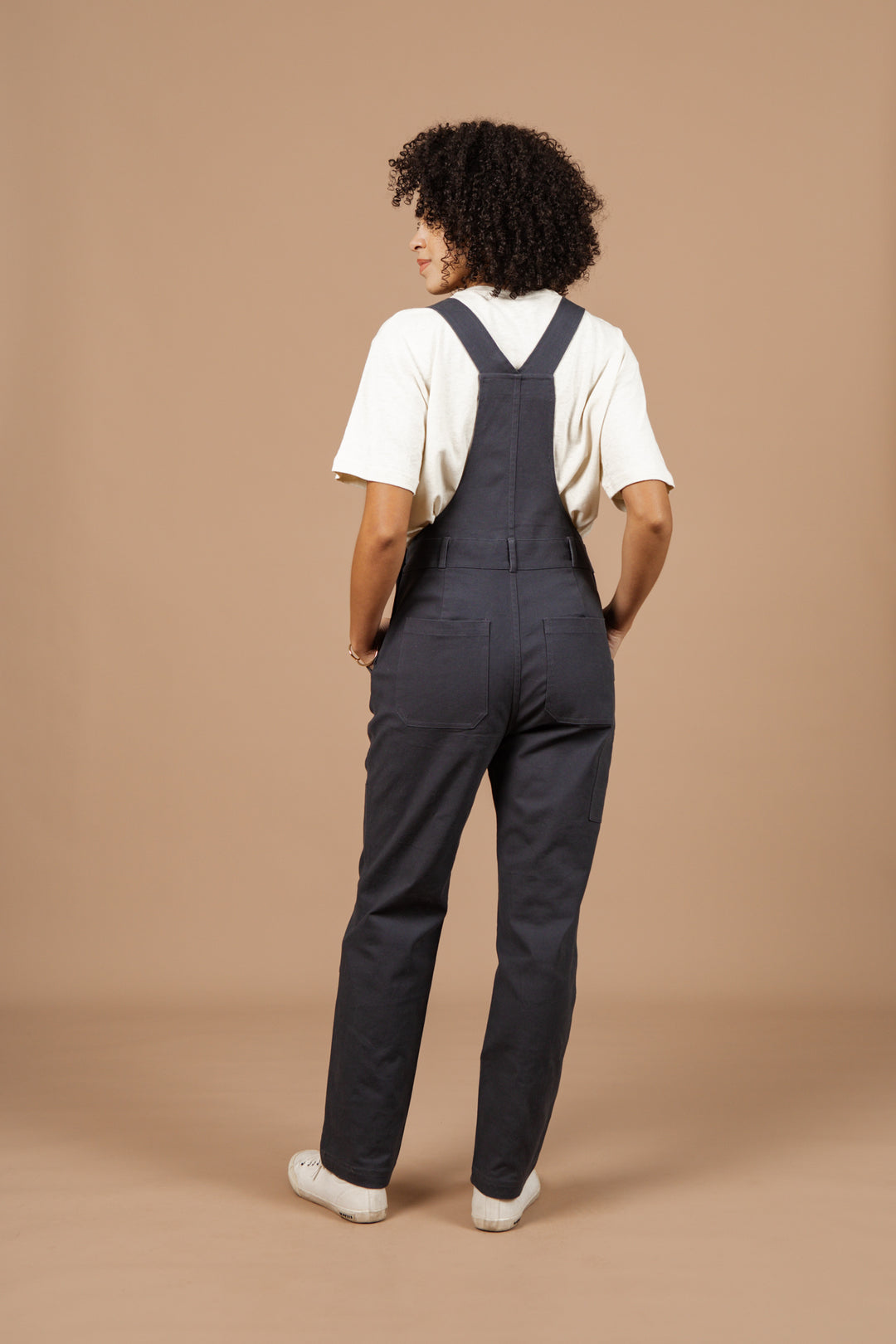 Person with curly hair faces away, wearing a white t-shirt, Bridge & Burn Tobin Utility Overall in dark charcoal canvas, and white sneakers against a plain beige background.