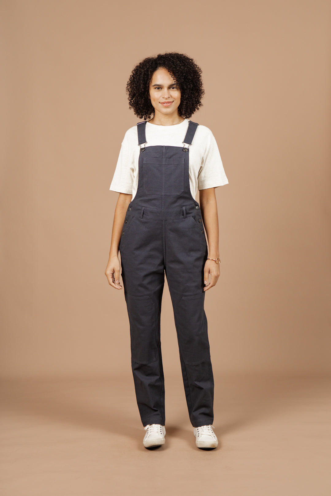 A person with curly hair stands against a beige background, wearing Bridge & Burn's Tobin Utility Overall in Dark Charcoal Canvas over a white t-shirt and white sneakers, smiling slightly with arms relaxed by their sides.